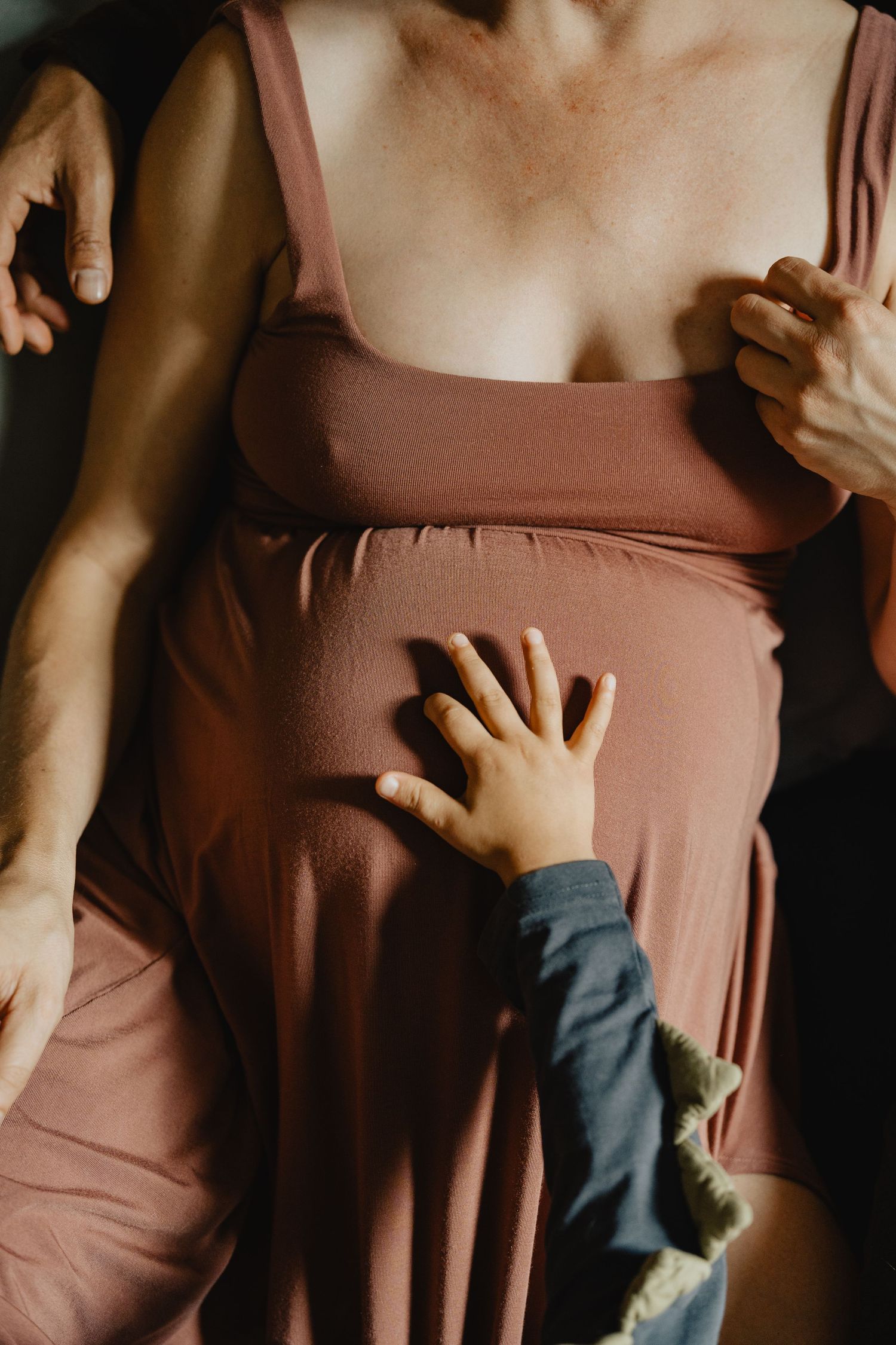 A small child's hand rests tenderly on a pregnant belly wearing a dusty rose colored dress.