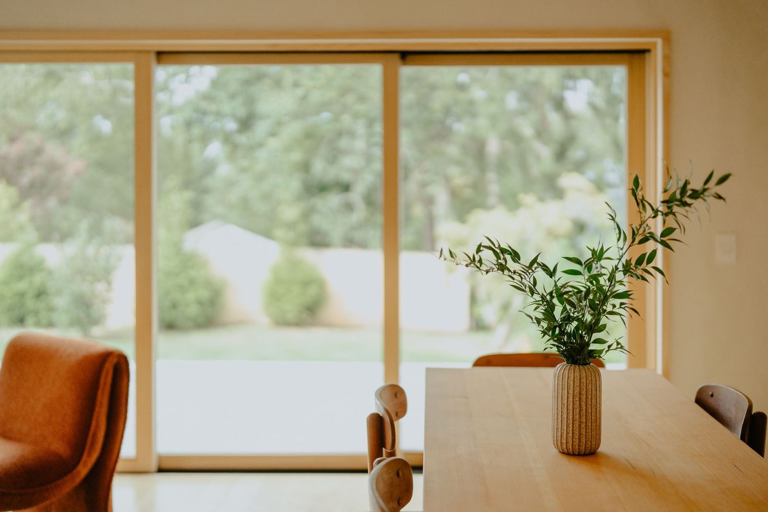 Bright dining area with wooden table and orange accent chair by large windows.