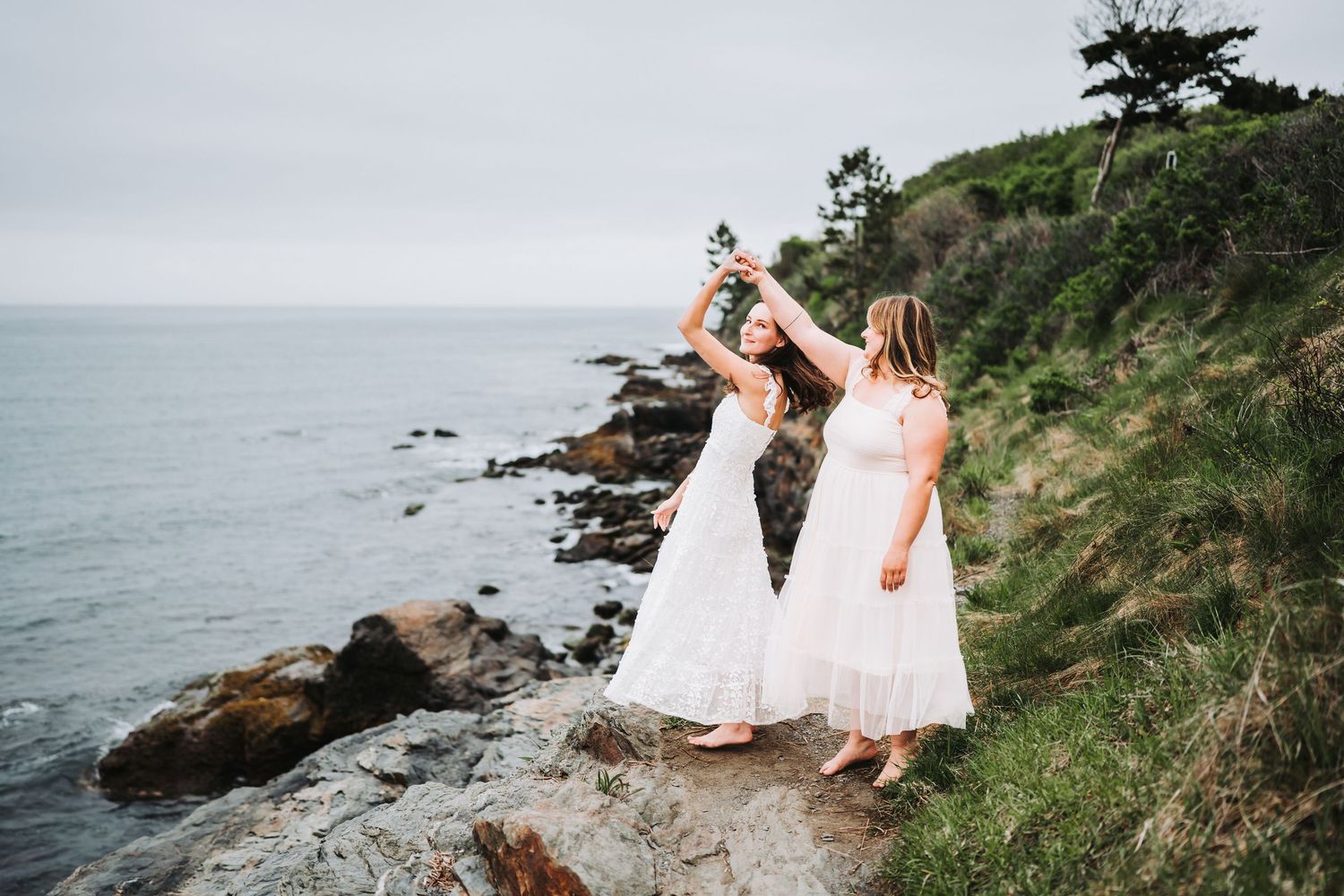 Two people in white dresses celebrate on a scenic coastal cliff overlooking the rocky Pacific Ocean shoreline.
