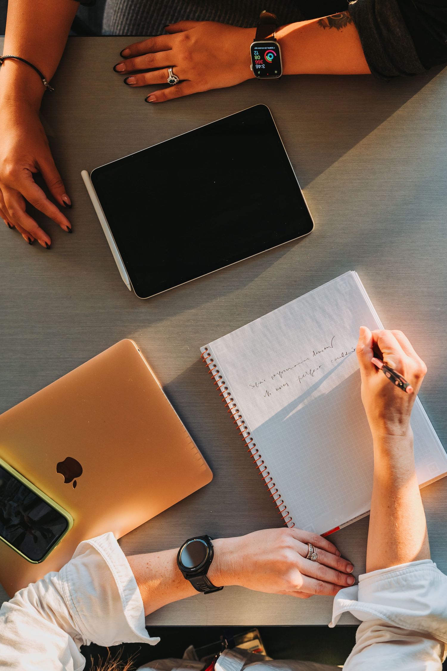 Overhead view of hands working at desk with Apple laptop, iPad, smartwatch and notebook for planning and note taking.