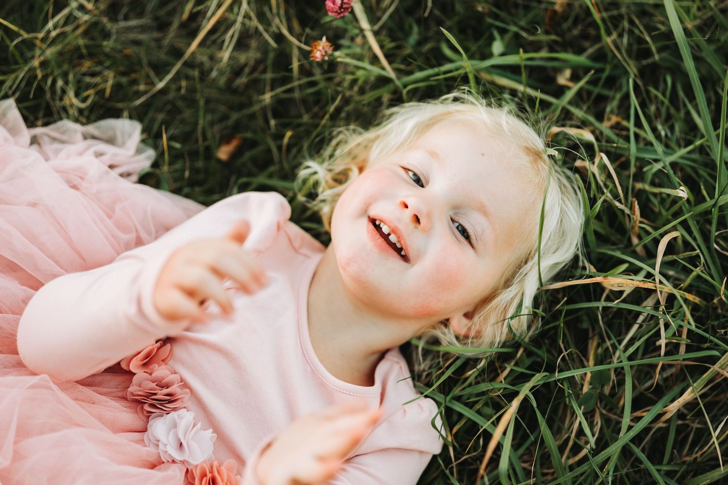 Child in pink dress laughing while laying in green grass.