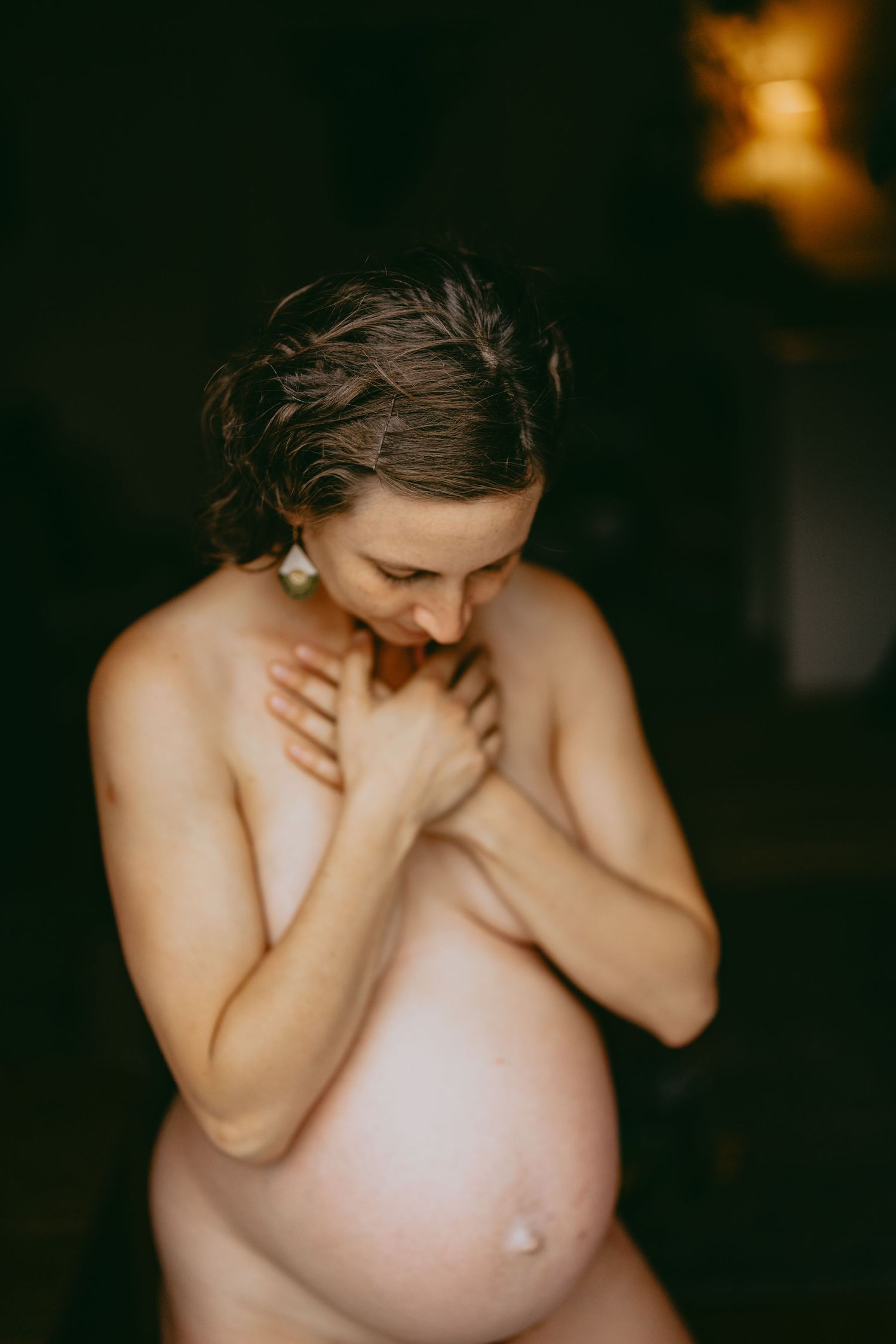 Artistic maternity photo showing pregnant woman embracing herself in dark, moody lighting.