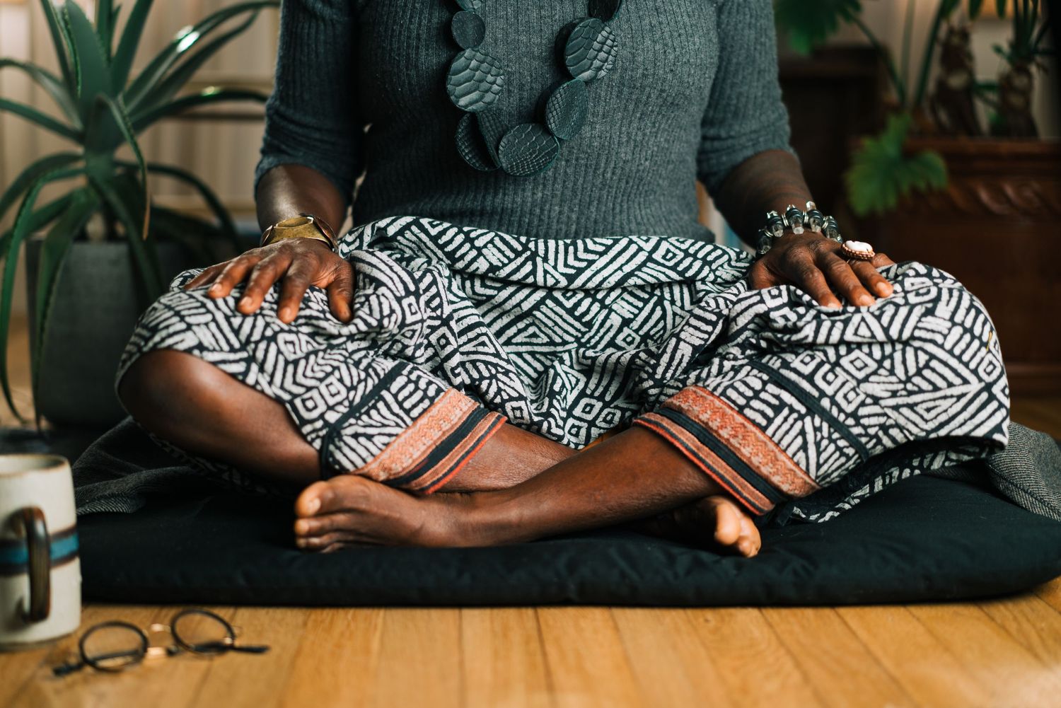 Person in meditation pose wearing patterned skirt with indoor plants in background.