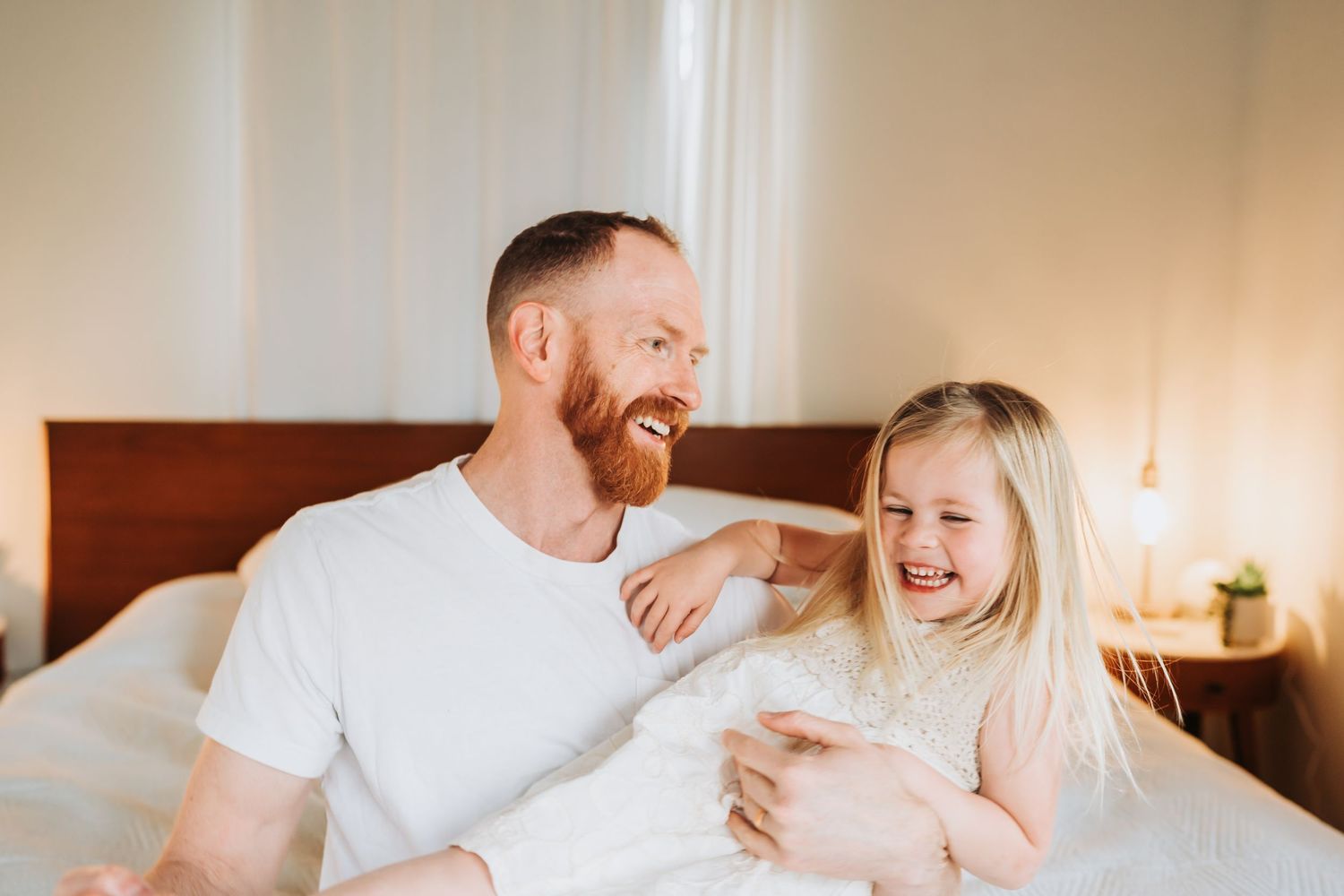 Laughing red-bearded father playing with blonde daughter on bed in bright cozy bedroom.