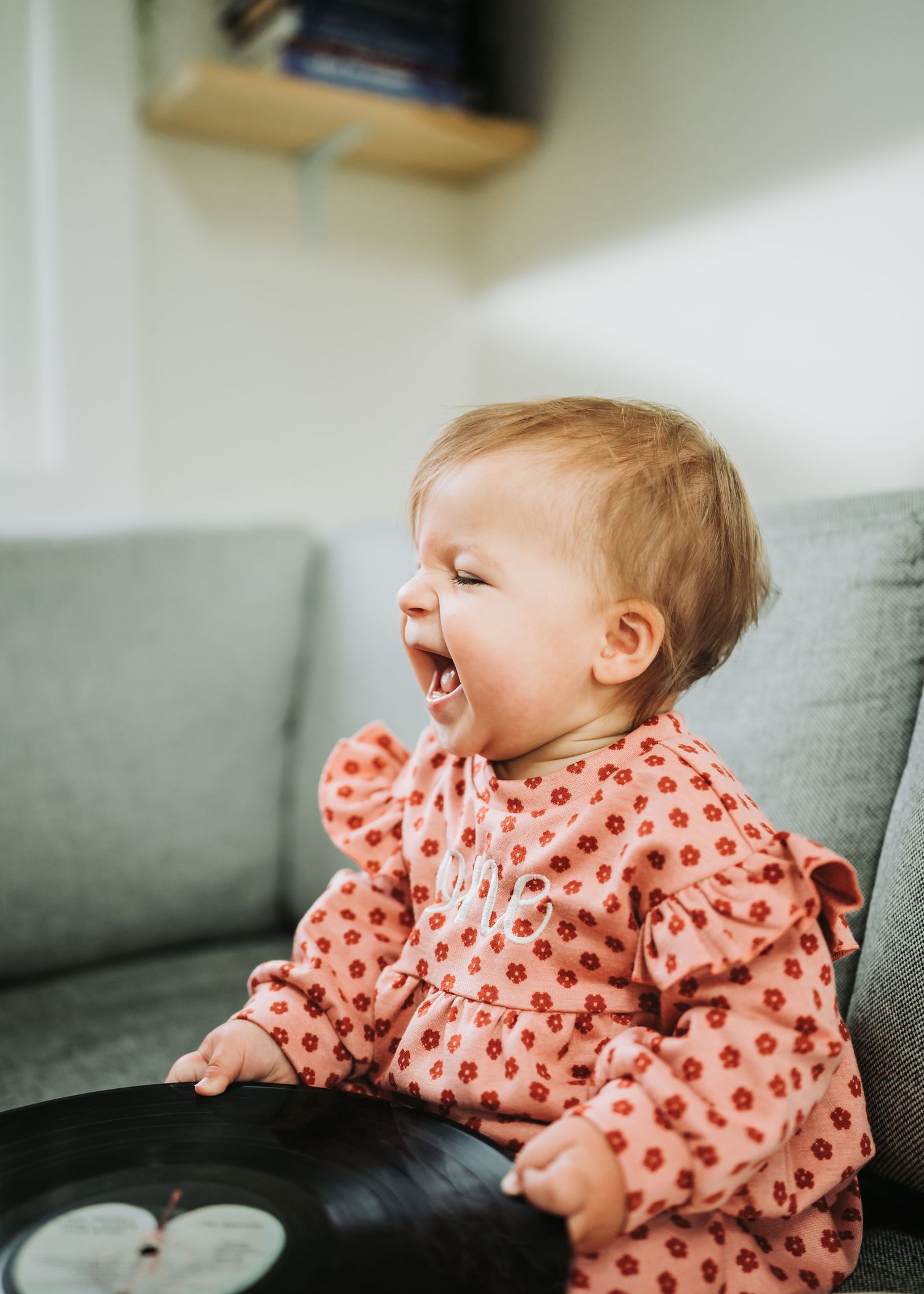 A baby in a coral patterned outfit laughs while sitting on a gray couch with dark cushions.