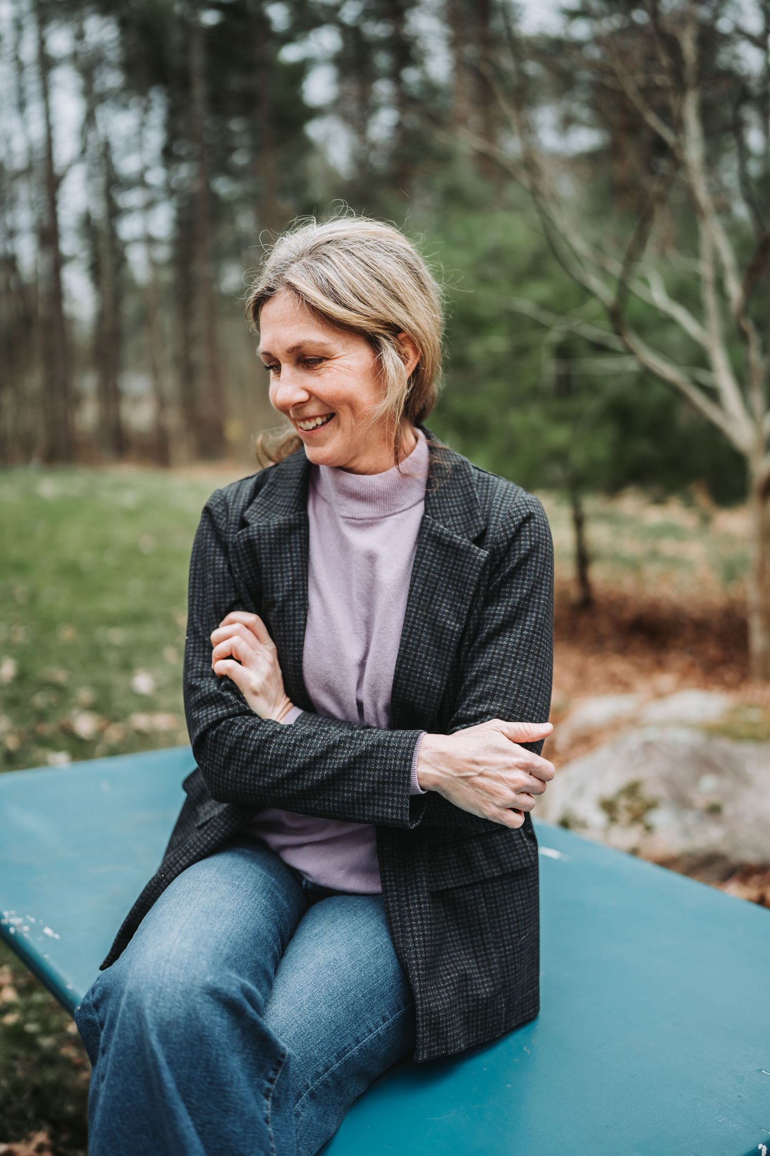 A person wearing a dark blazer and jeans sits on a blue surface outdoors in a wooded setting.
