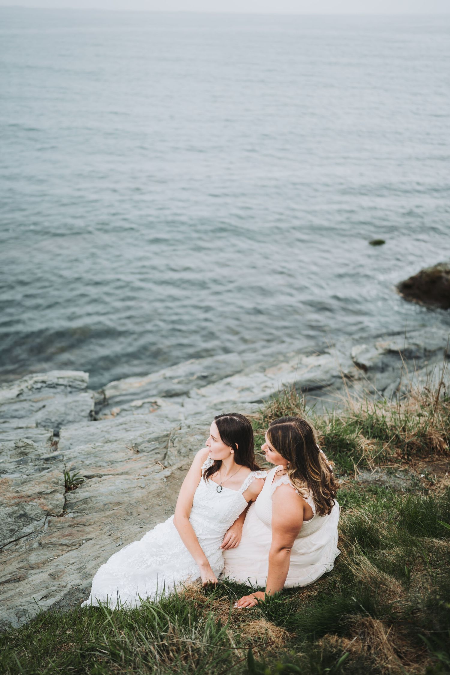 Two people in white dresses lay together on a coastal cliff overlooking a misty ocean.