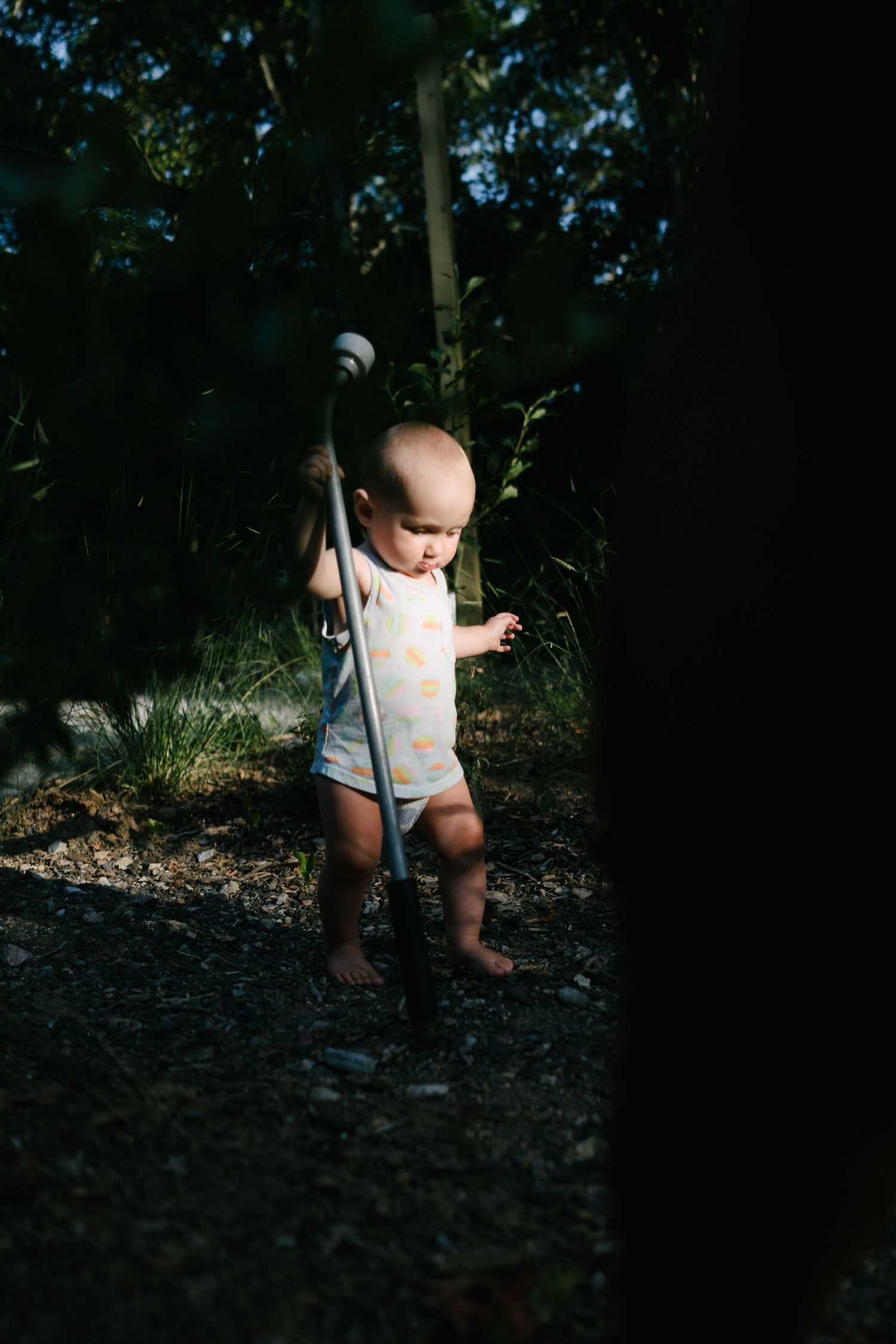 A toddler in a white onesie exploring outdoors in dappled sunlight among trees.