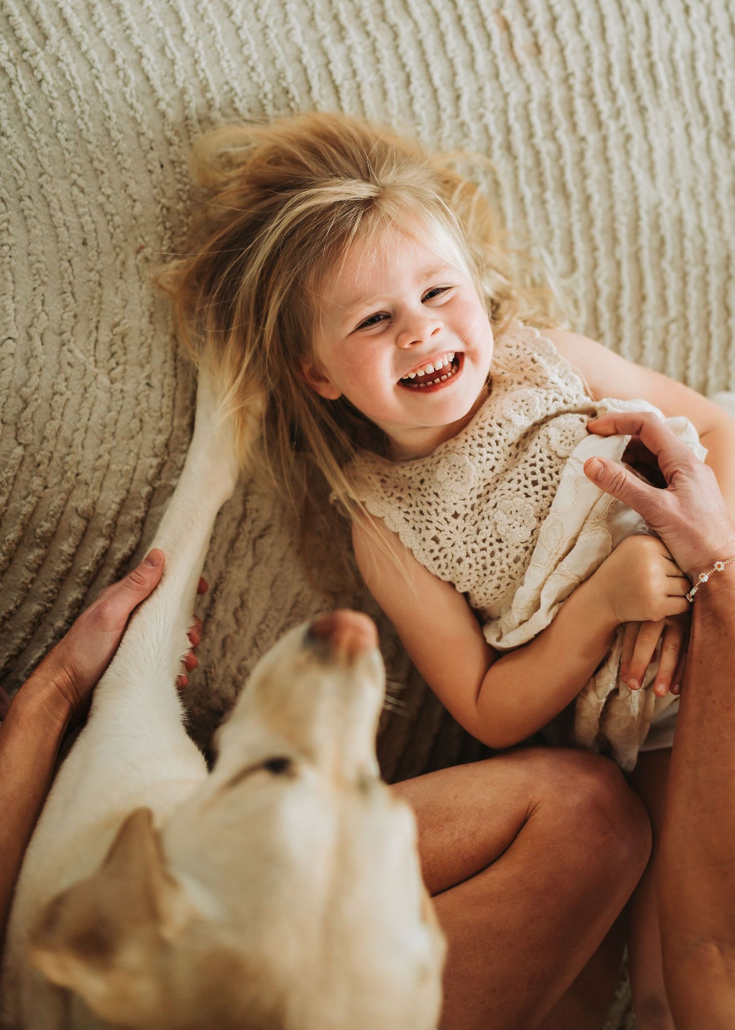 Happy blonde toddler girl laughing joyfully while playing with a yellow Labrador dog on a cozy blanket.