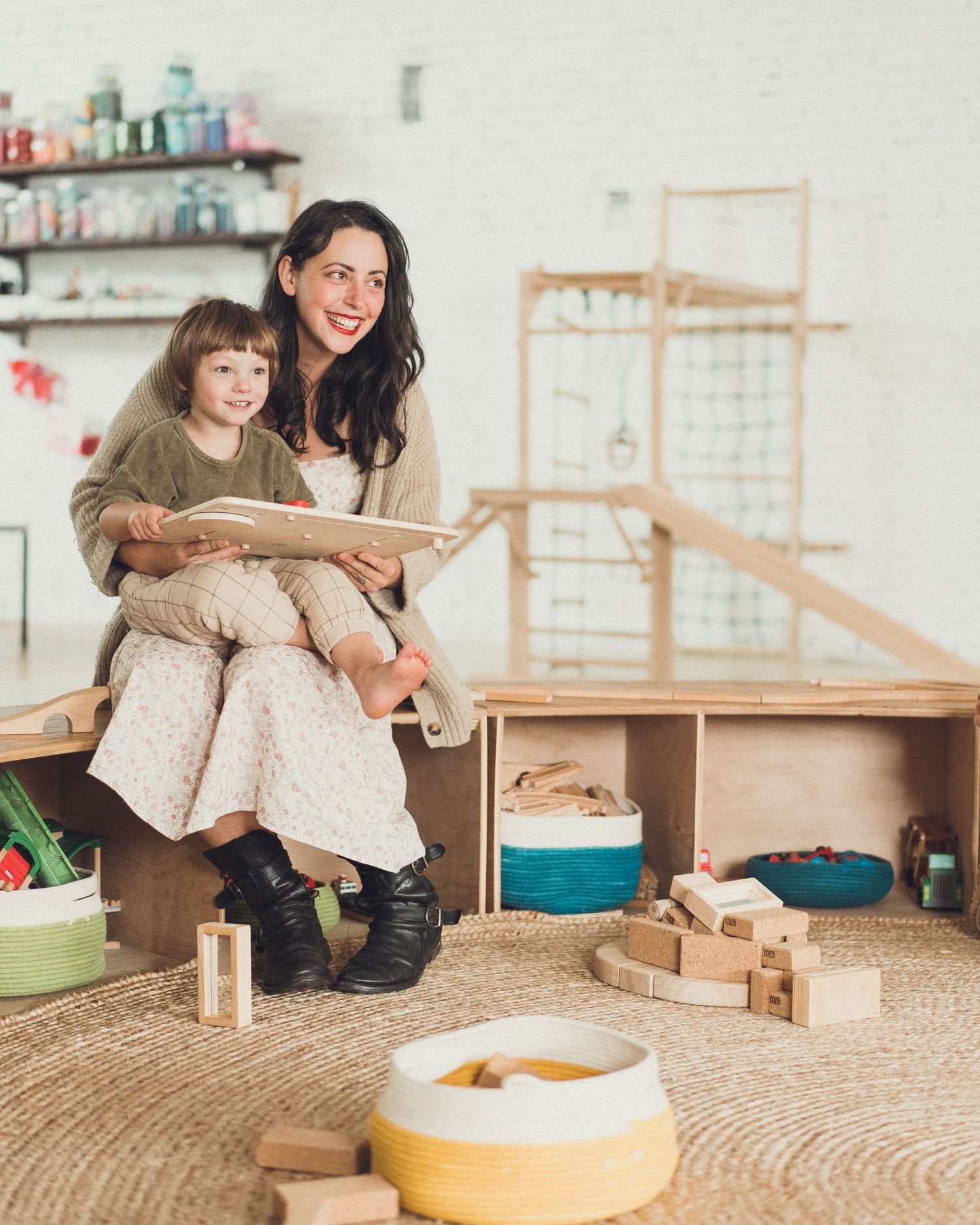 Adult and child read together in cozy playroom surrounded by wooden toys and storage bins.