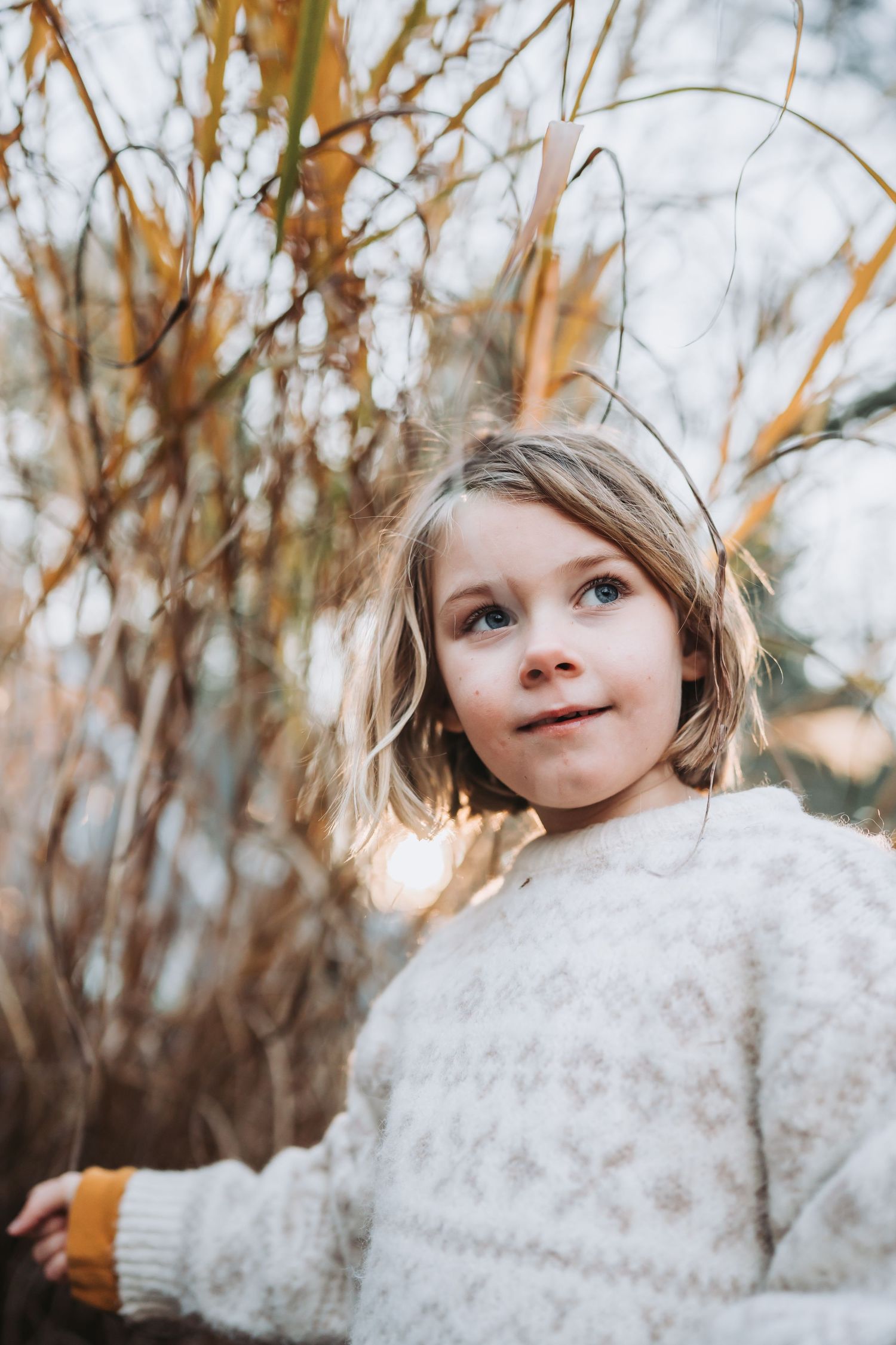 Child peeking out from behind fluffy white snow with autumnal trees in the background during a winter photoshoot.