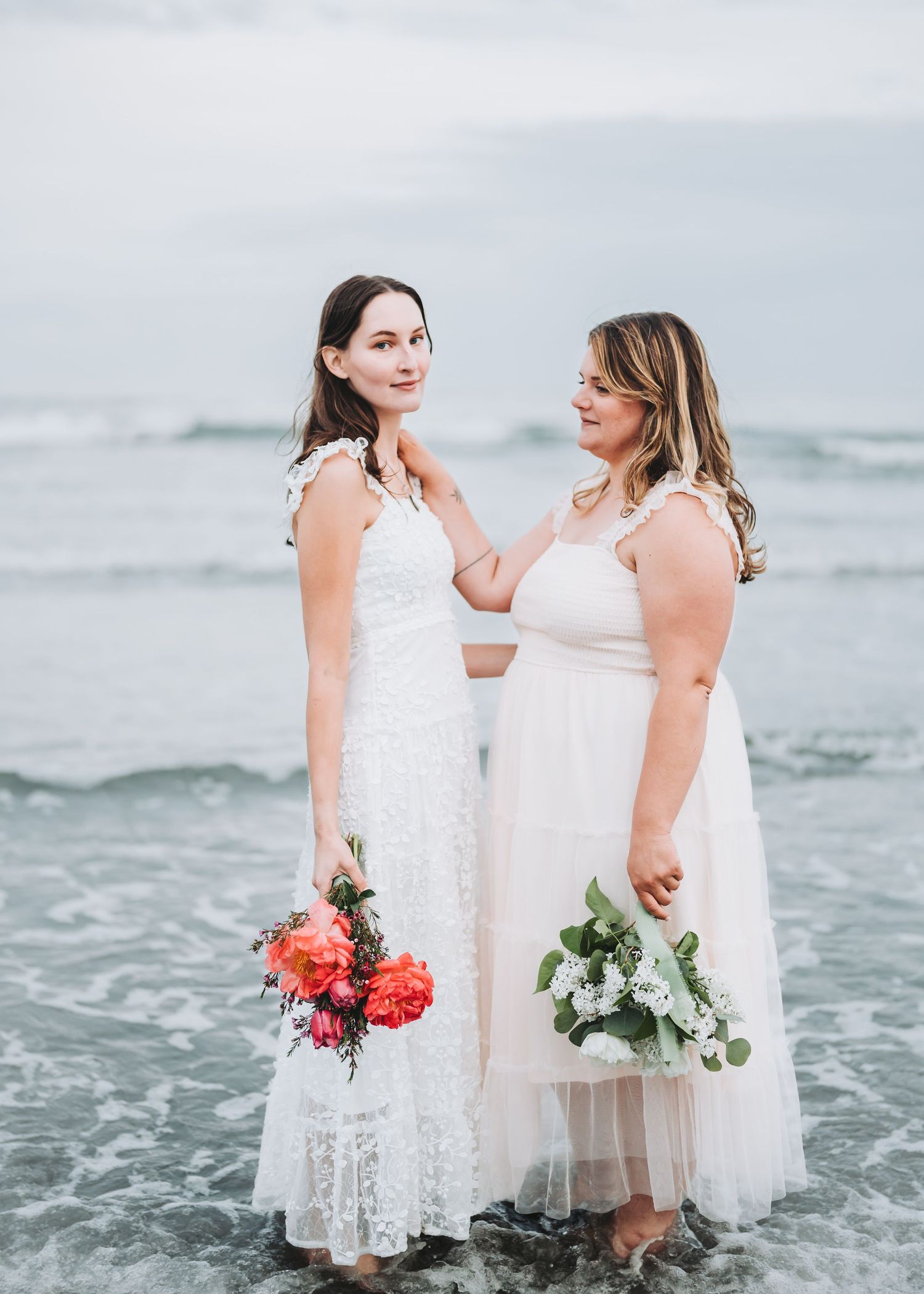 Two people in white dresses stand together at the beach, holding bouquets while waves crash behind them.