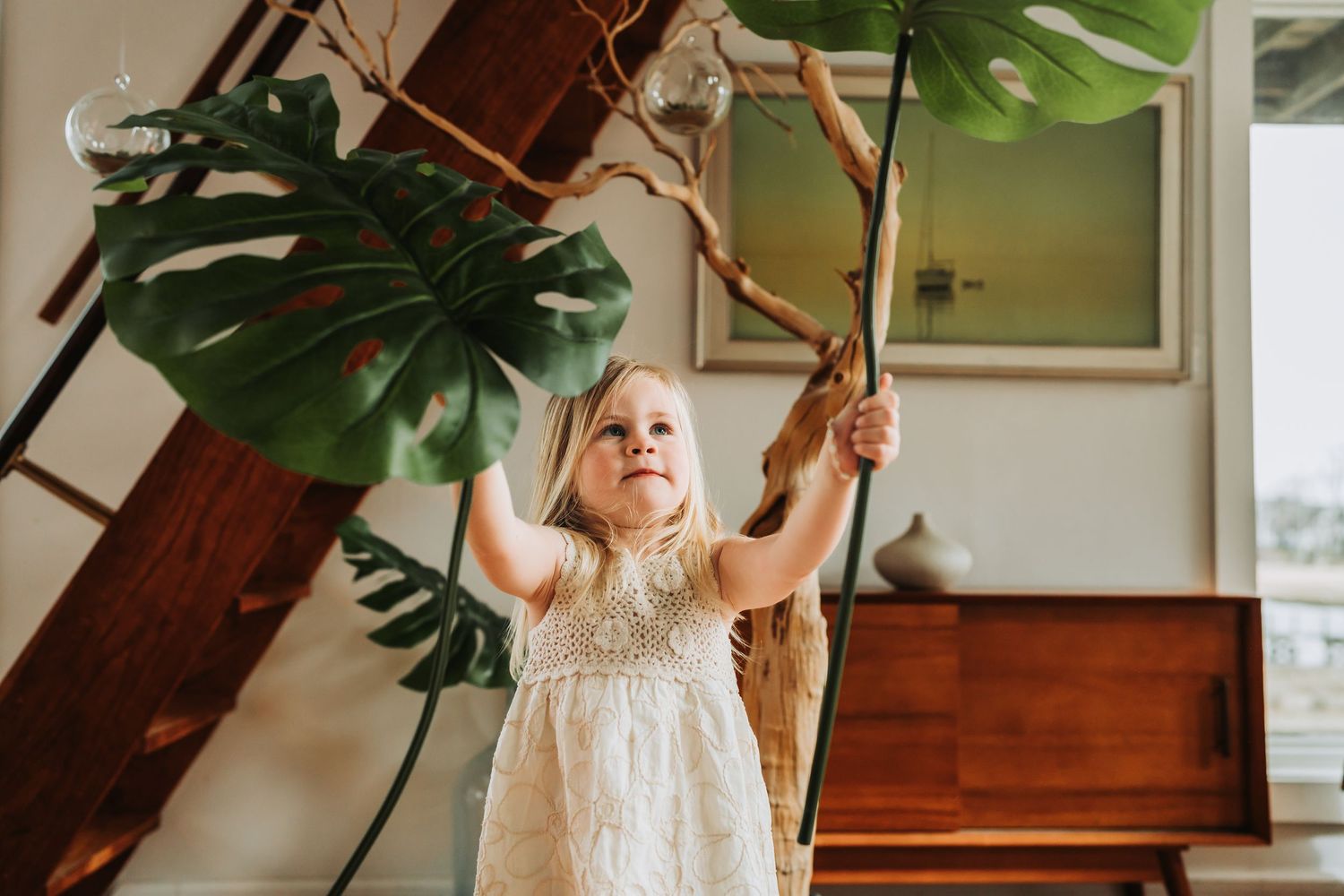Toddler girl in white dress reaching up to touch monstera leaves and branches indoors.