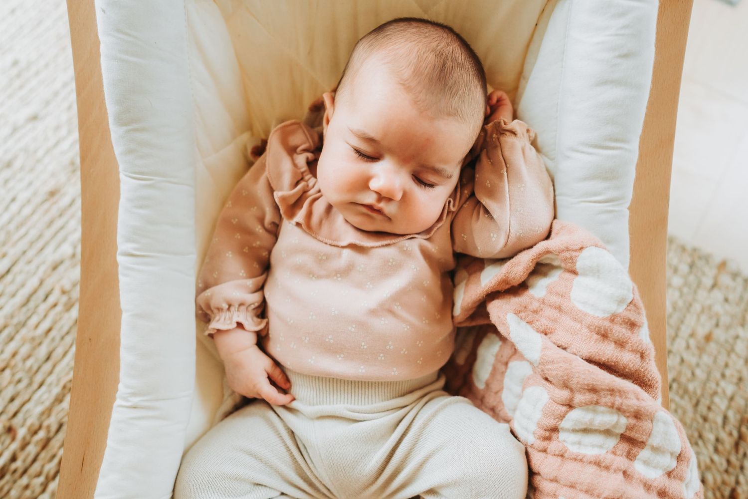Peaceful sleeping newborn in light colored outfit rests on white cushioned surface with pink blanket.