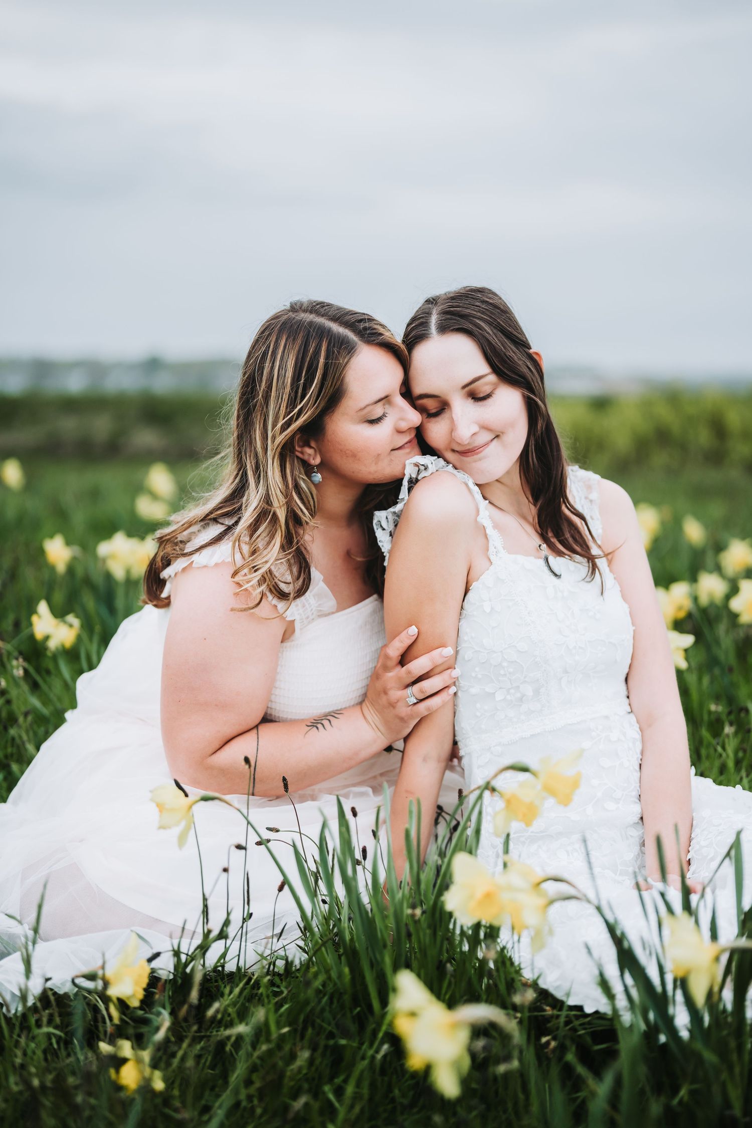 Two people in white dresses share tender moments while sitting in a field of yellow daffodils on a spring day.