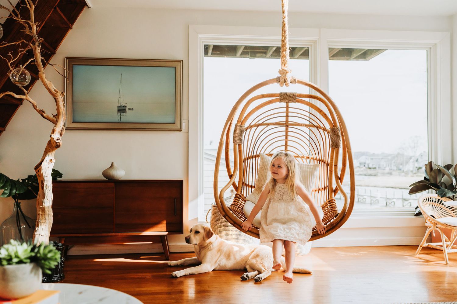 Young girl in white dress stands beside a golden retriever under a rattan hanging chair in a bright living room.