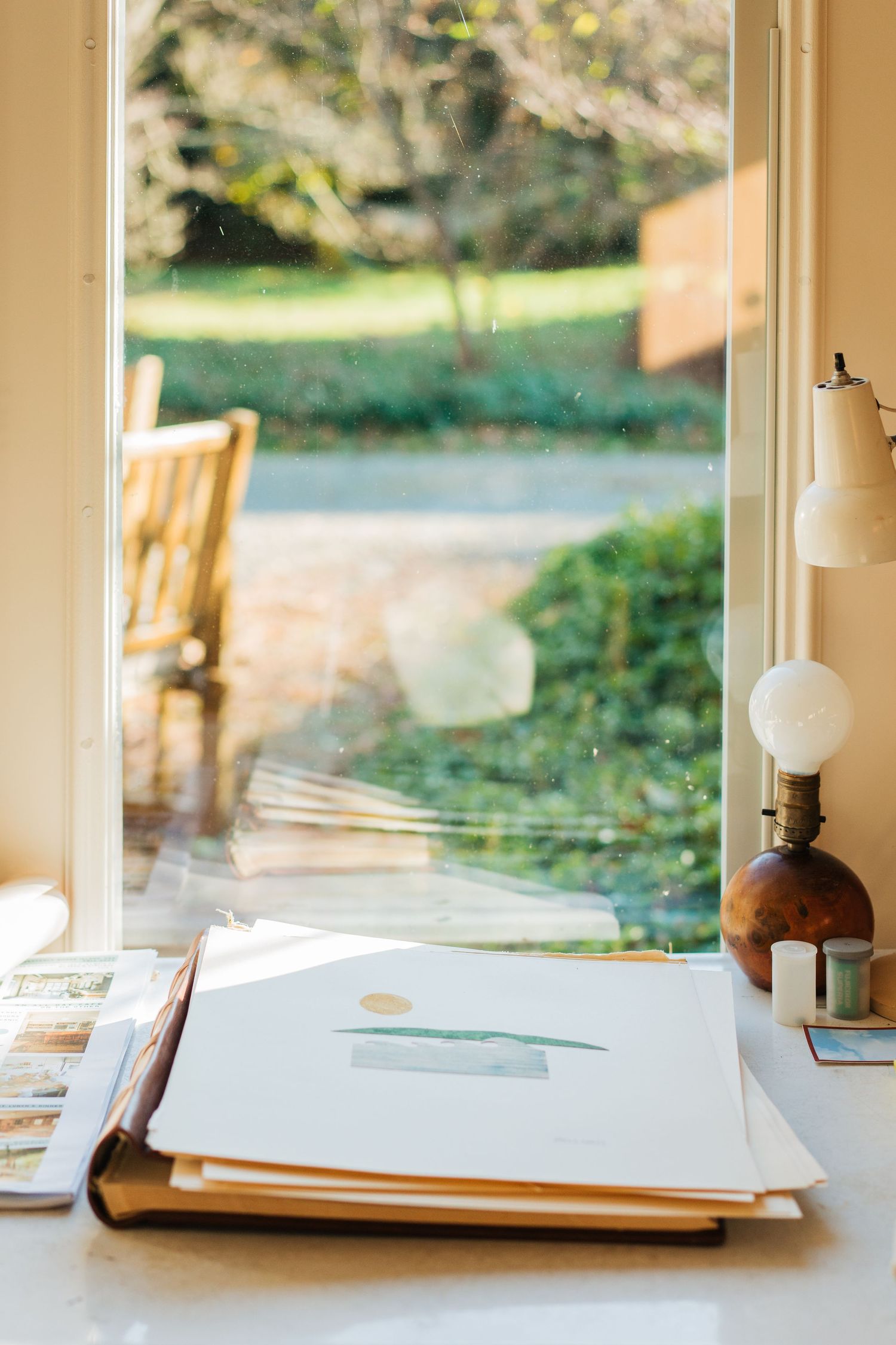 A desk with papers and an antique lamp next to a window overlooking a garden.