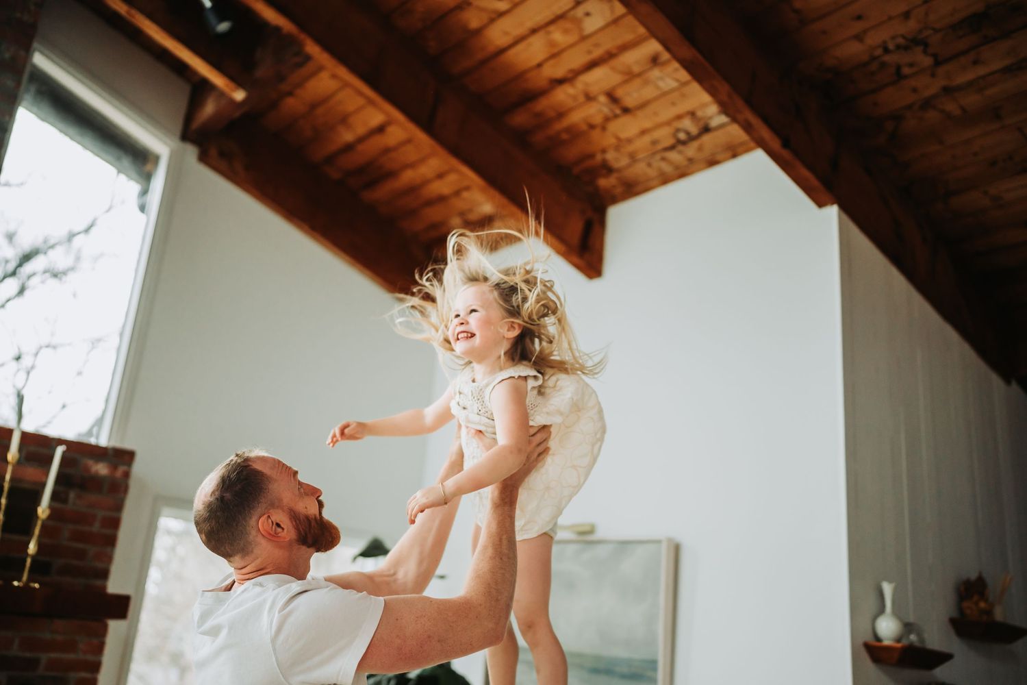 A bearded father joyfully lifts his laughing little girl in a bright, modern living room with wooden ceiling beams.