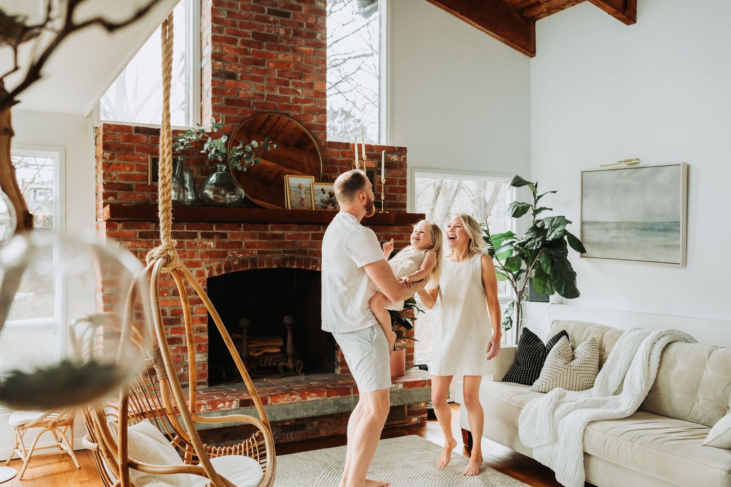 Family dancing in bright living room with brick fireplace, dad holding baby while mom smiles nearby.
