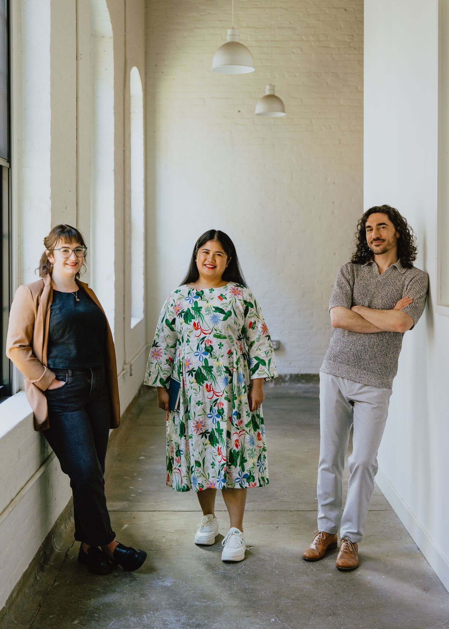 A group of people stand against a white wall in a hallway with pendant lights overhead.