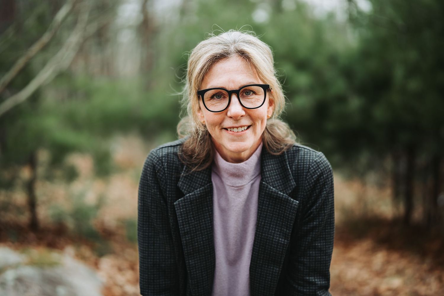 A smiling person in glasses wearing a black cardigan and lavender turtleneck stands against a blurred outdoor background.