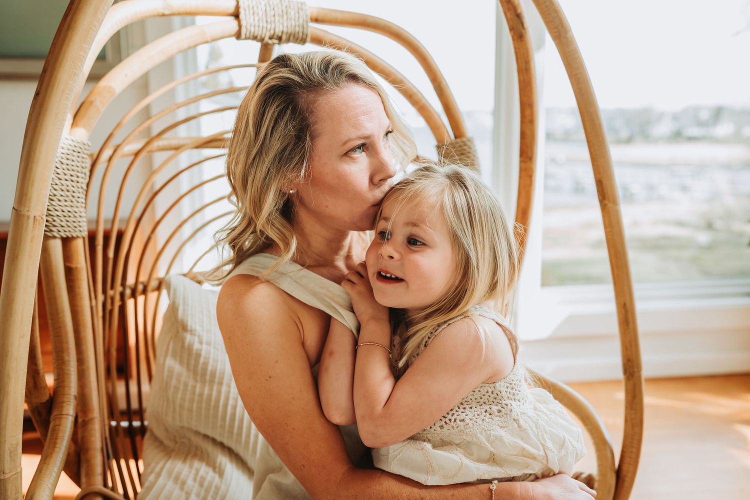 A mother kisses her young blonde daughter while sitting in a rattan chair by a bright window.