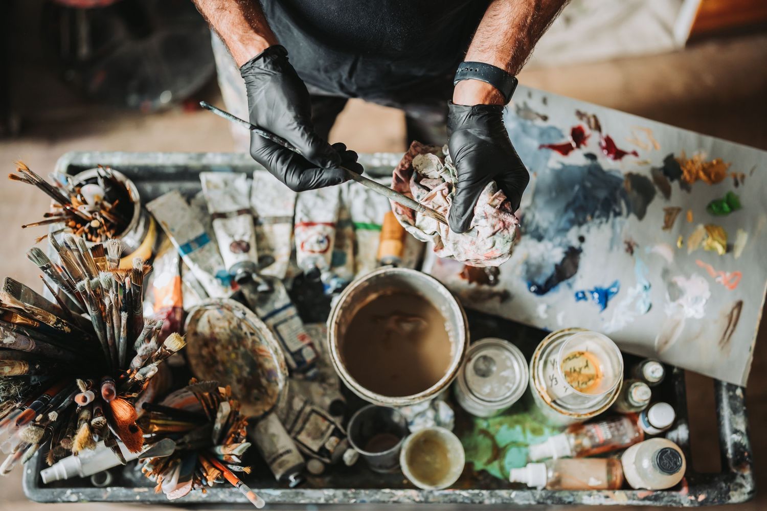 Artist's hands in black gloves work with paint and brushes at a messy art table covered in supplies and paint jars.