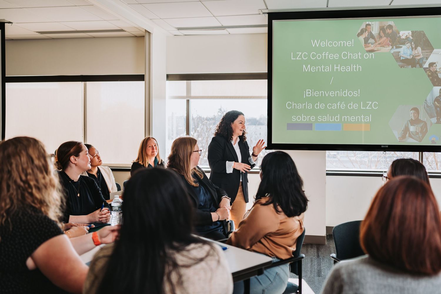 Business professionals gather in a modern conference room for a presentation with green slides projected on screen.