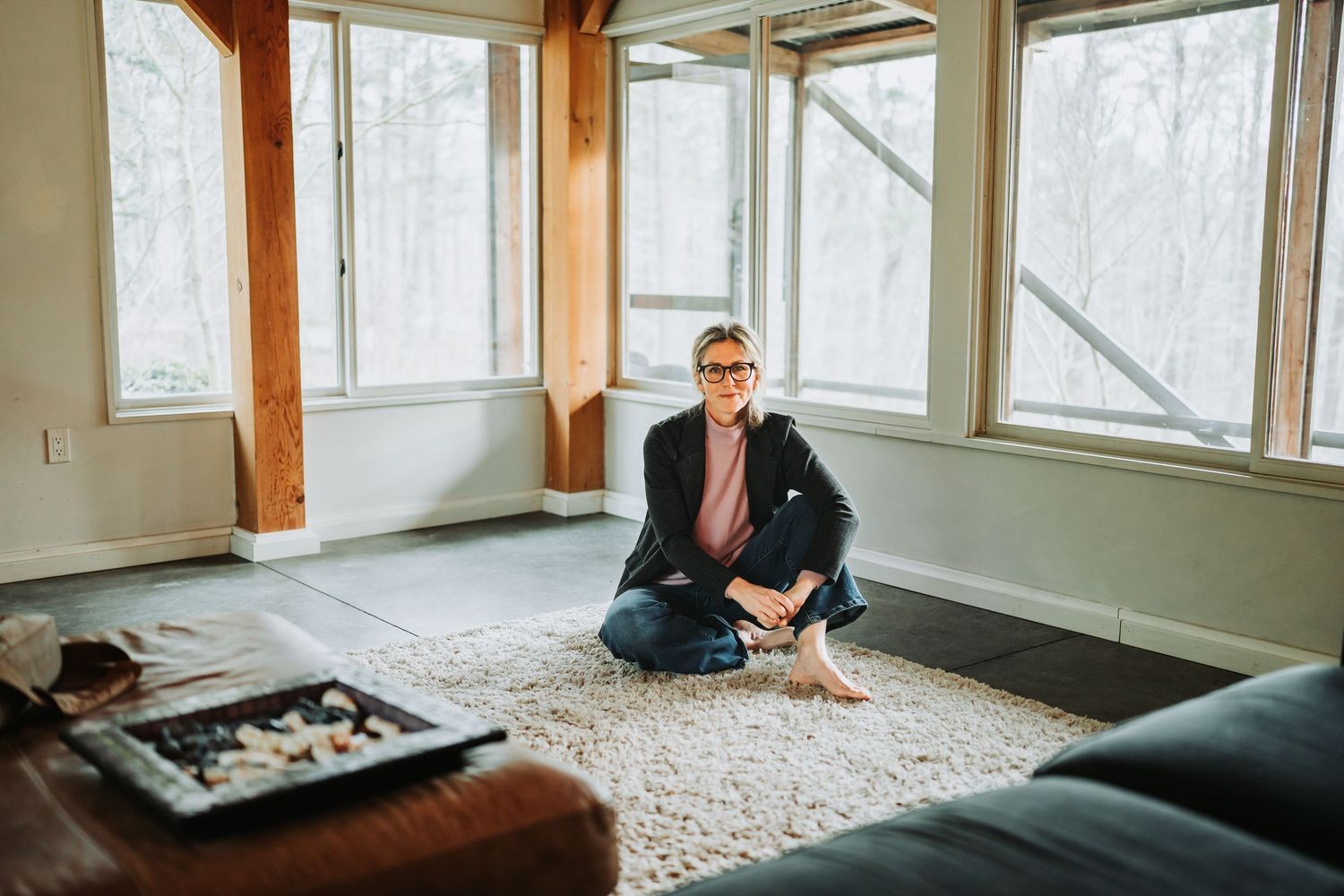 A person sits on a bench or daybed near large windows in a sunlit modern room with wooden beams.