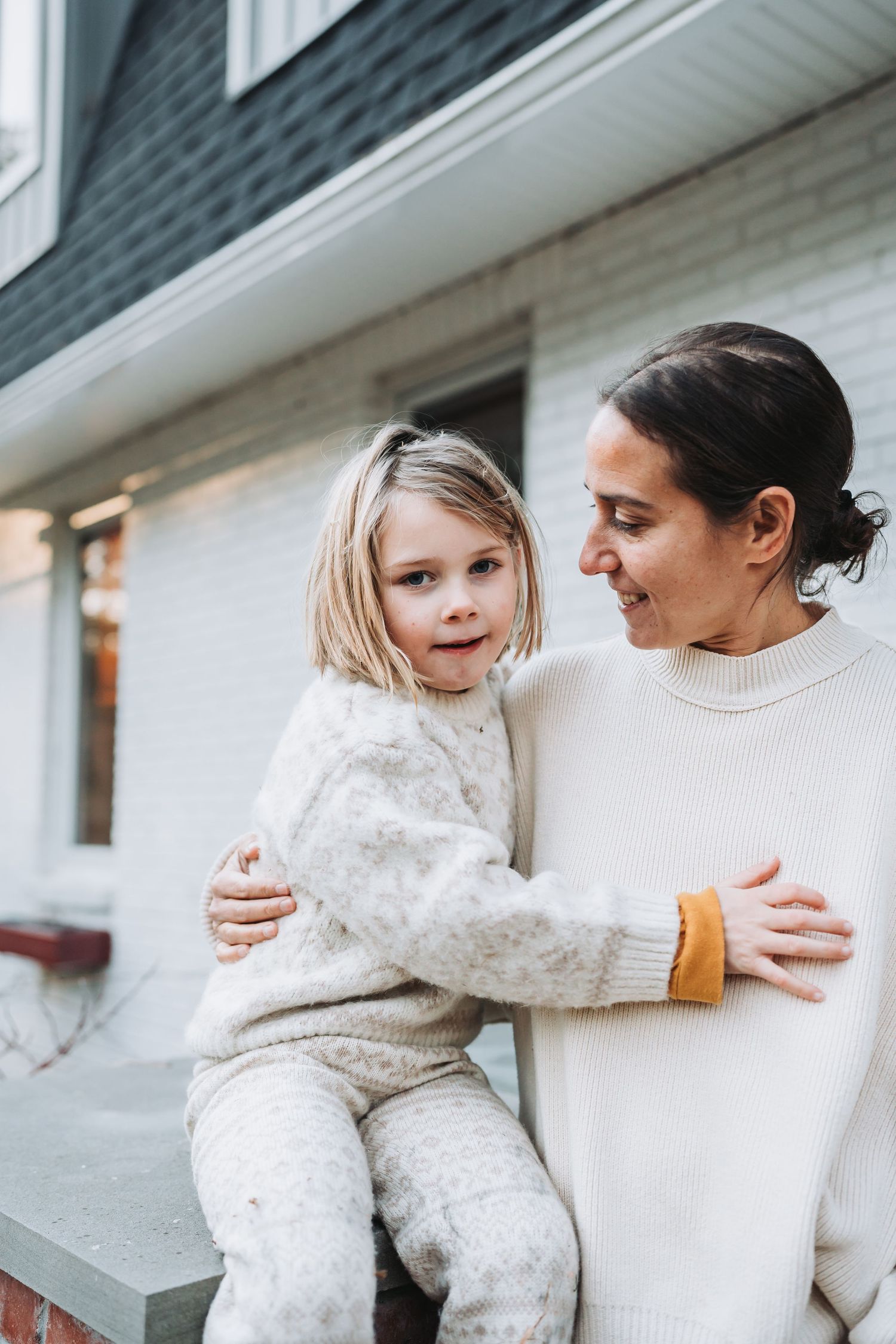 Parent and child wearing cozy cream-colored sweaters share tender moments on outdoor porch steps.