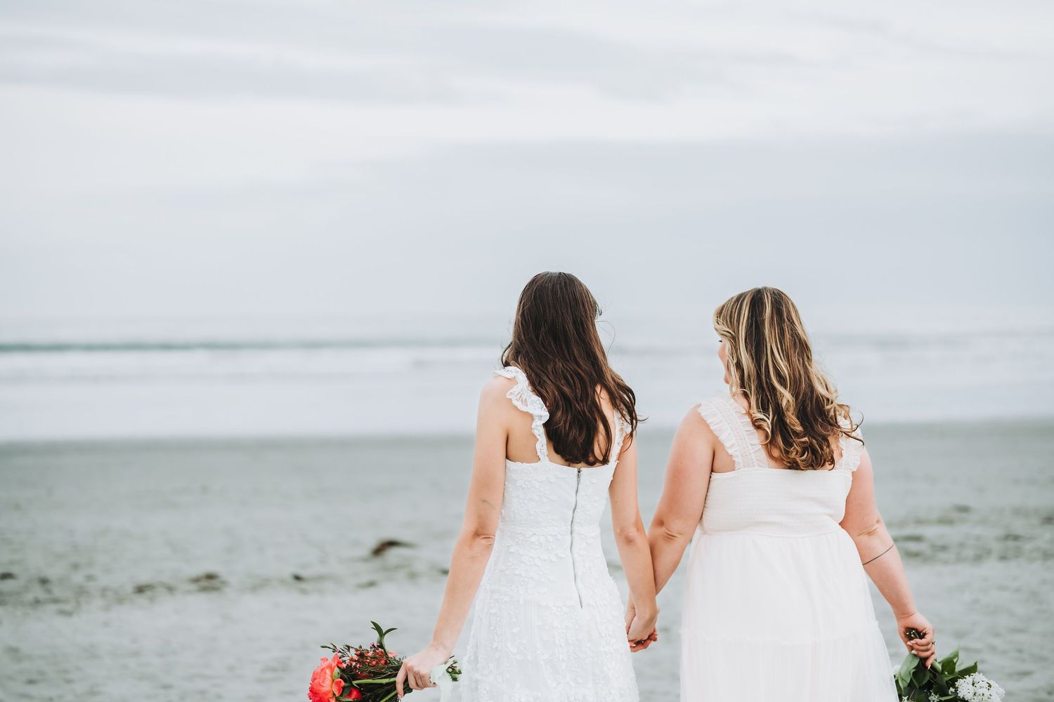 Two people in white dresses walking along a scenic beach holding bouquets on their wedding day.