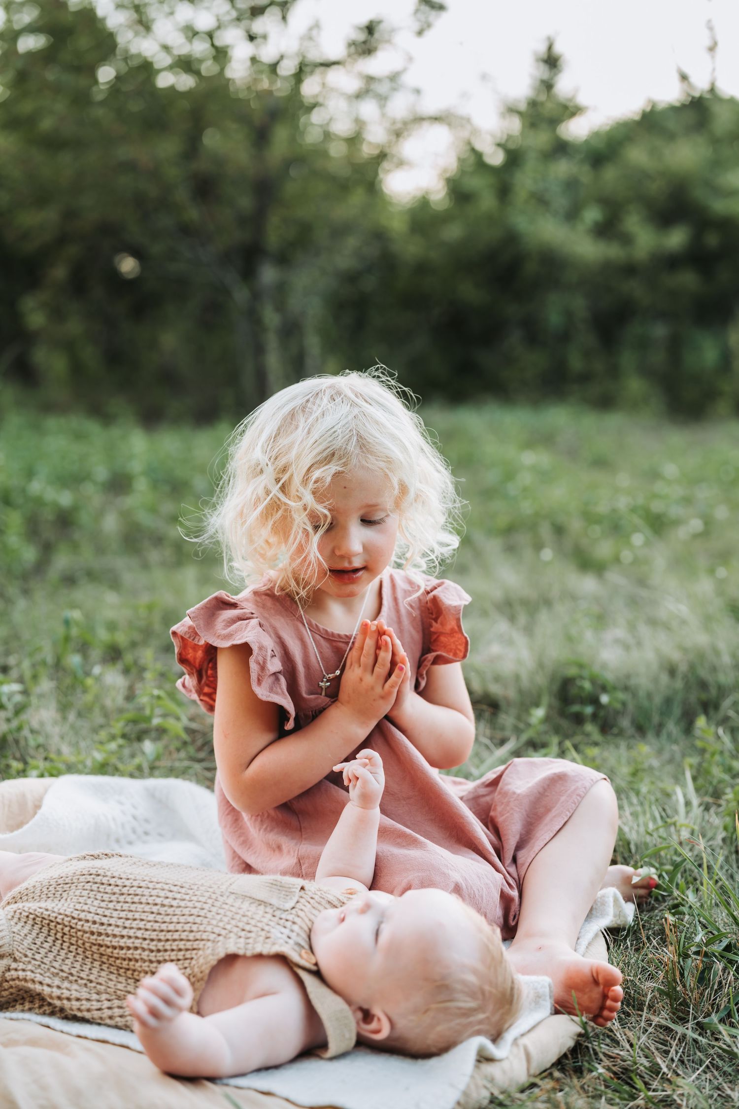 Little one in pink dress sitting on blanket in grass examining something with curiosity in a sunlit garden.