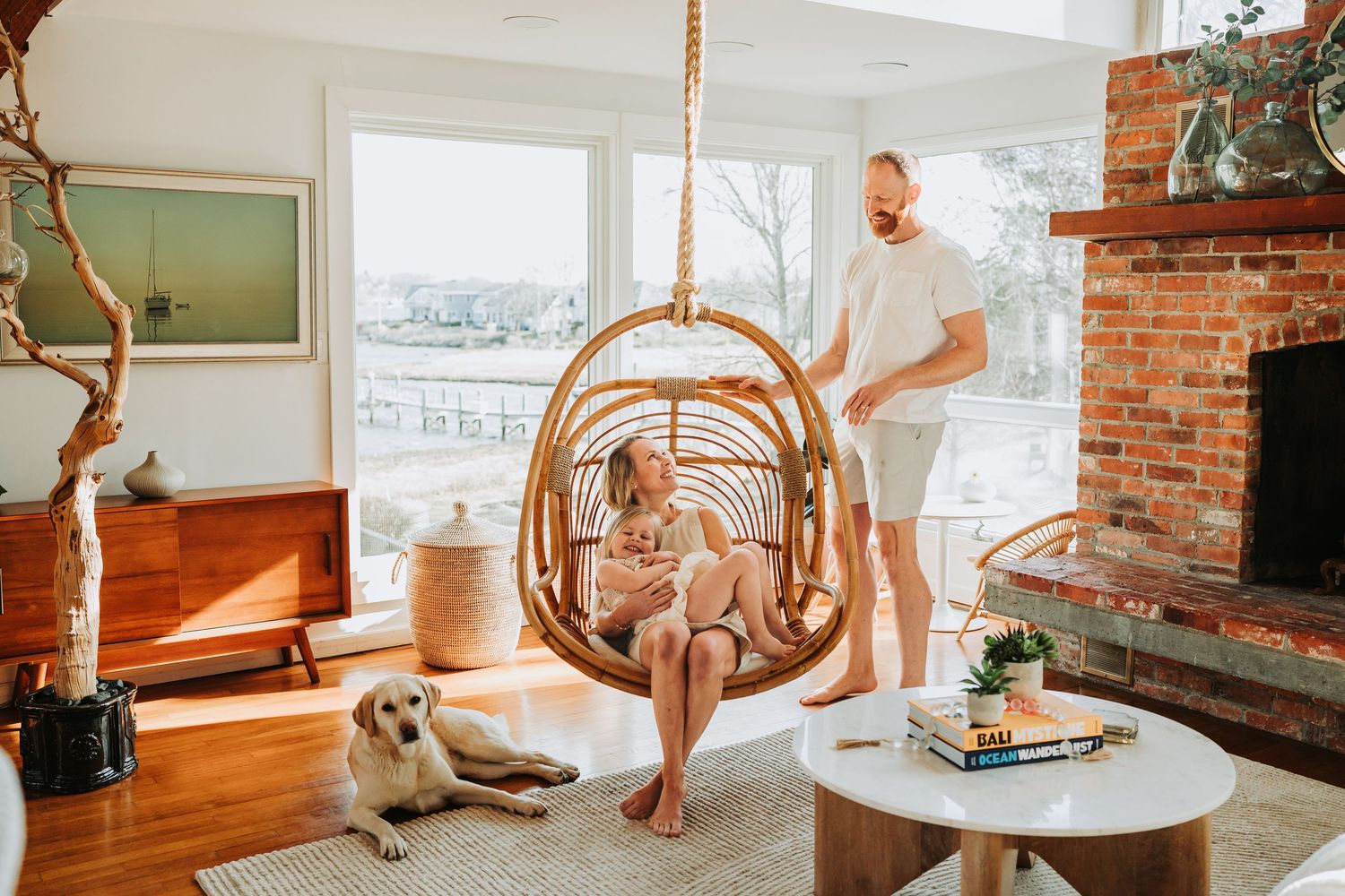 Couple relaxing in boho living room with rattan swing chair, golden retriever, and brick fireplace.