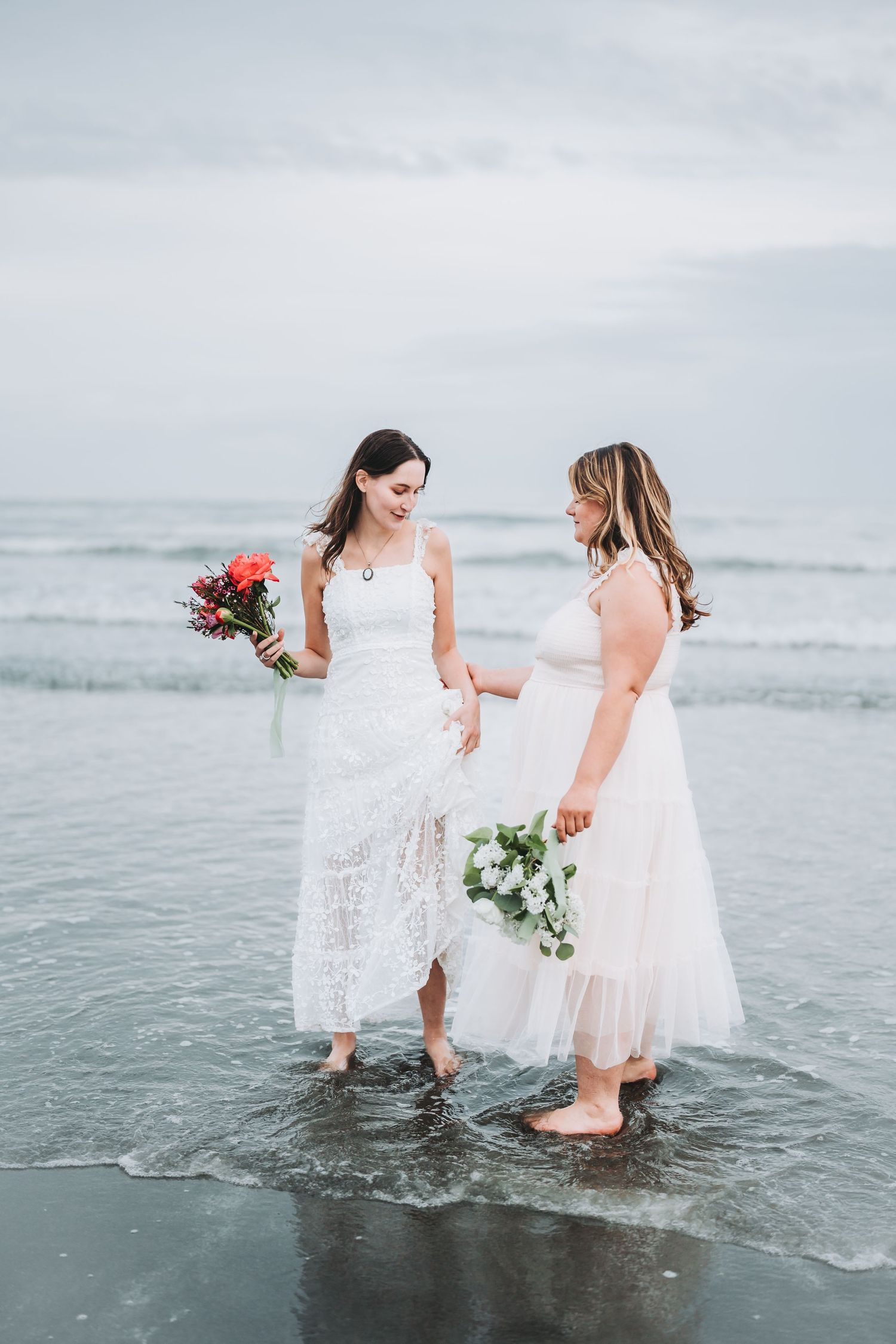 Two women in white dresses wade into the ocean water while holding bouquets at sunset.