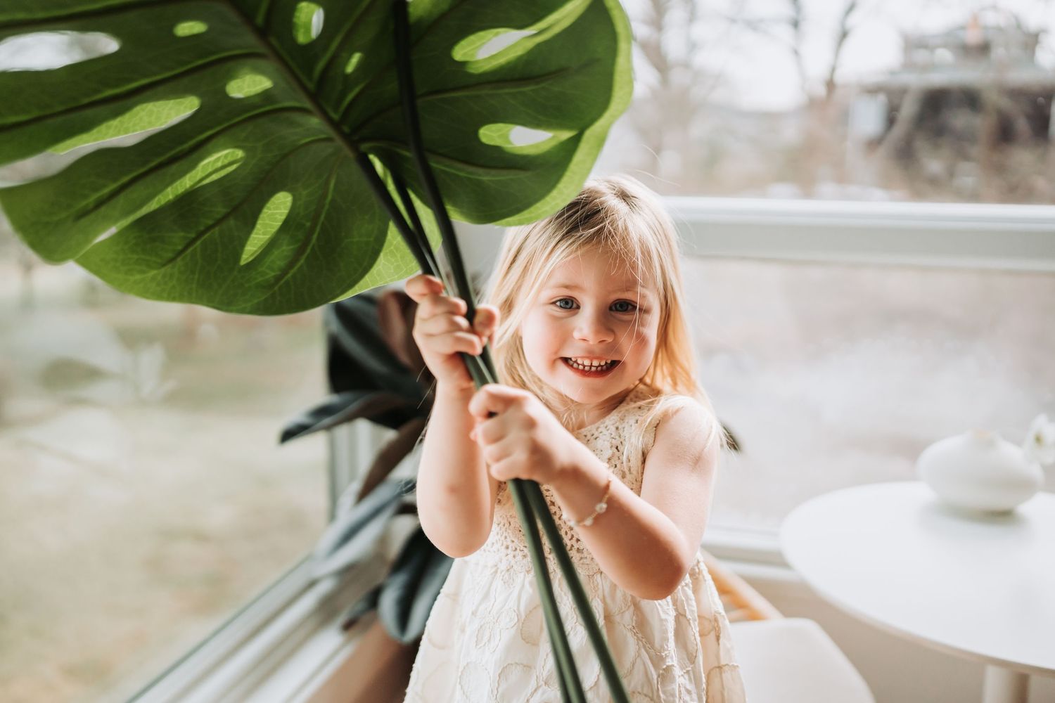 Happy toddler girl in white dress hugs a large monstera leaf plant near a bright window indoors.