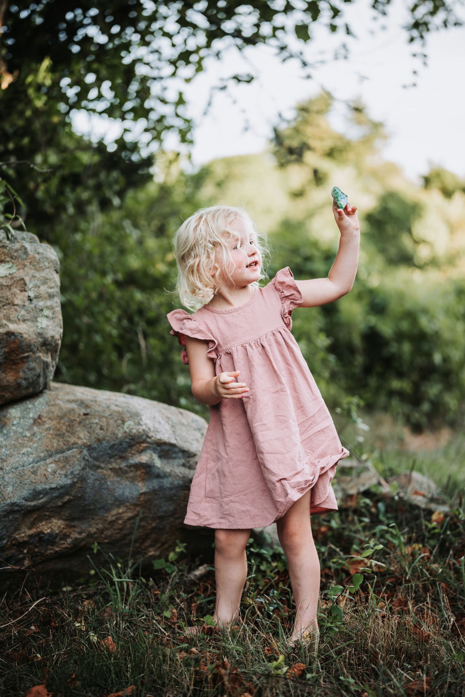A small child in a pink dress explores a rocky outdoor area while playing in nature during golden hour.