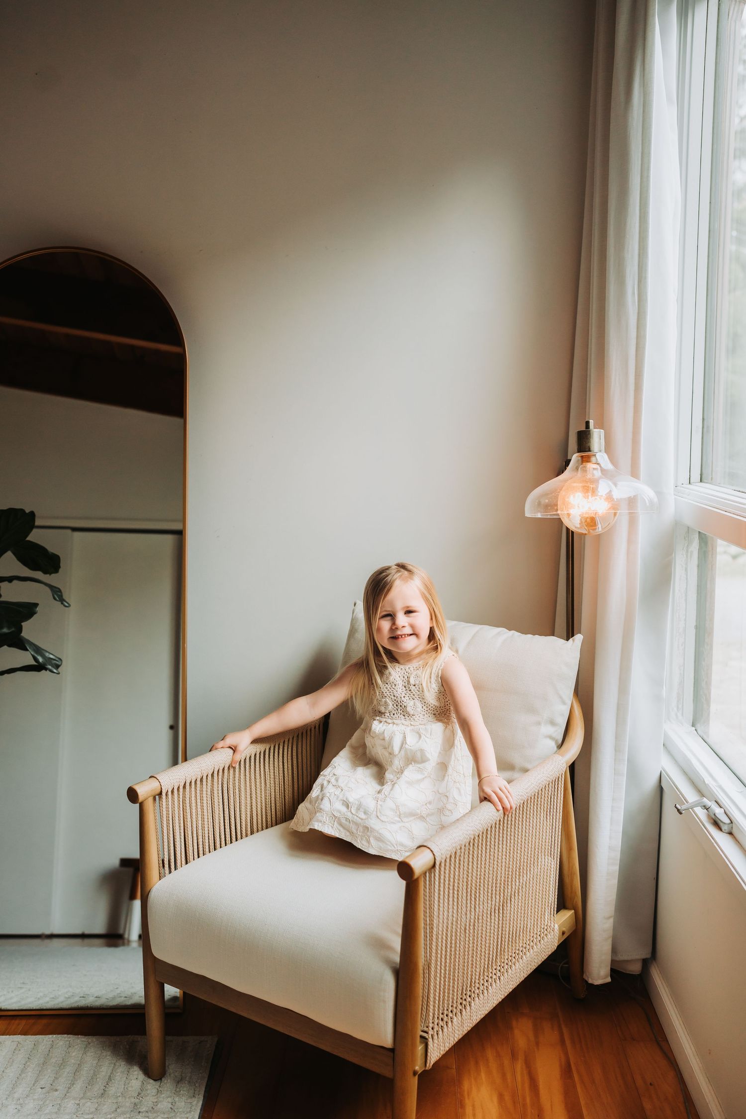 A smiling young girl in a white dress sits in a rattan armchair near a bright window in a cozy room.