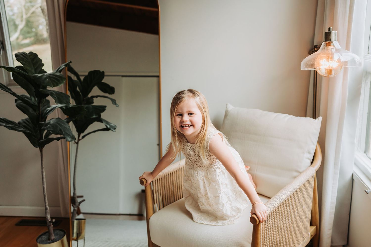 Happy toddler girl in white dress standing in modern cream armchair beside indoor fiddle leaf fig tree.
