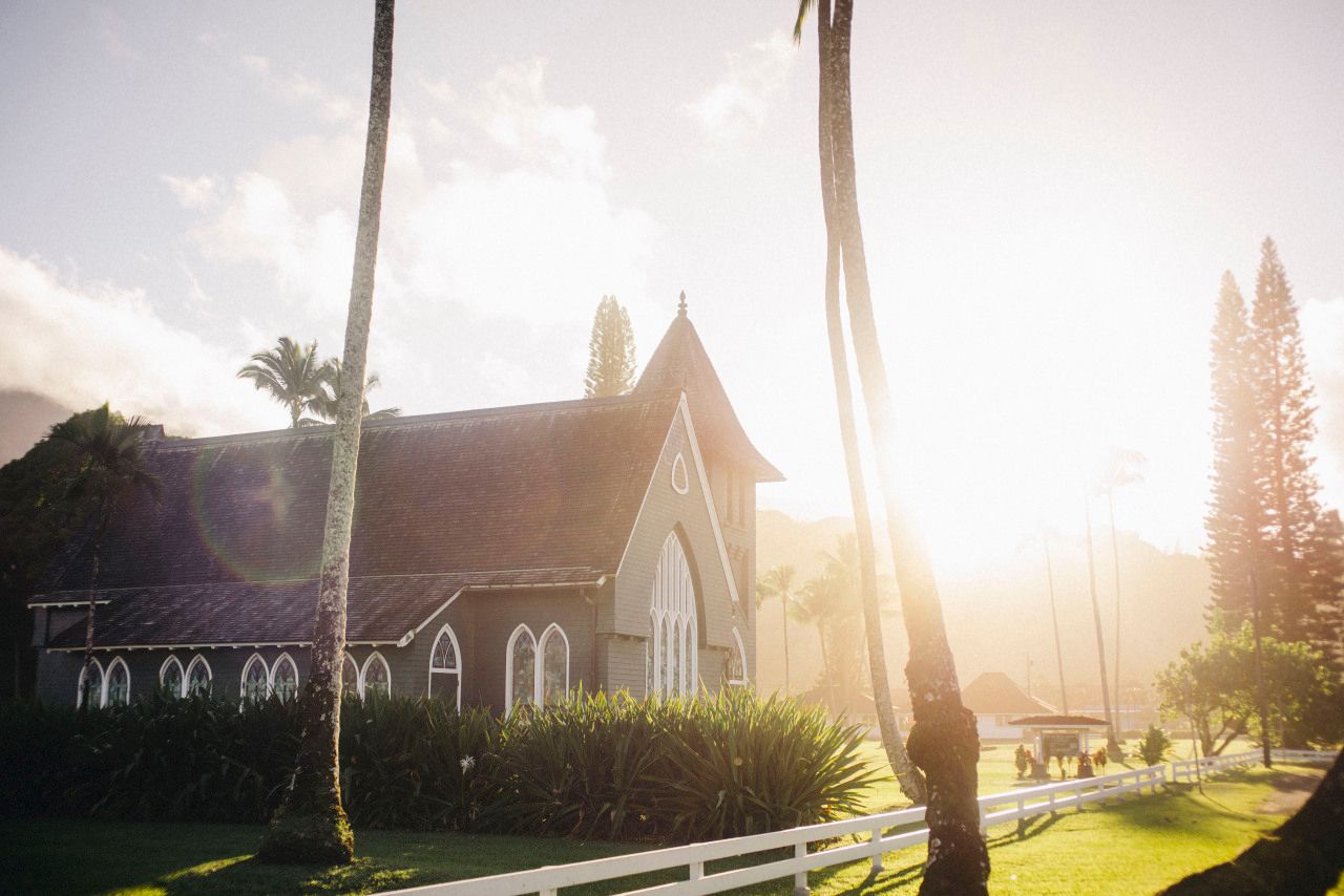 Golden sunlight streams through trees beside a historic stone church building with arched windows and steeples.