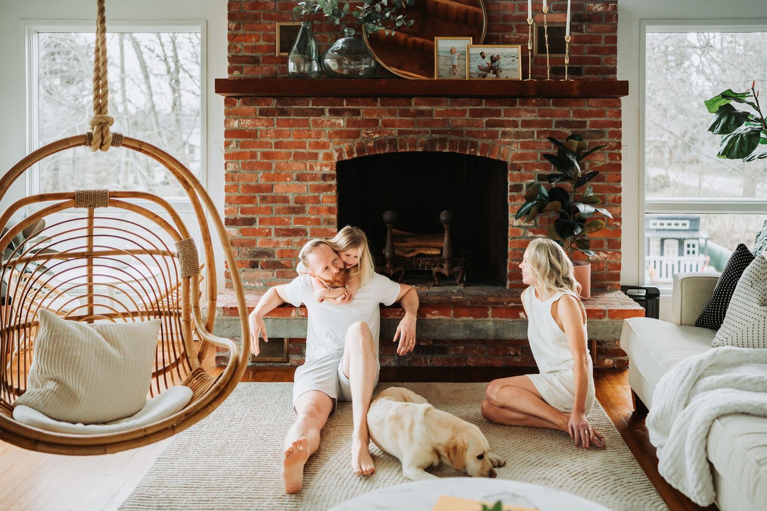 Family with dog relaxing on living room floor near brick fireplace with hanging rattan chair.