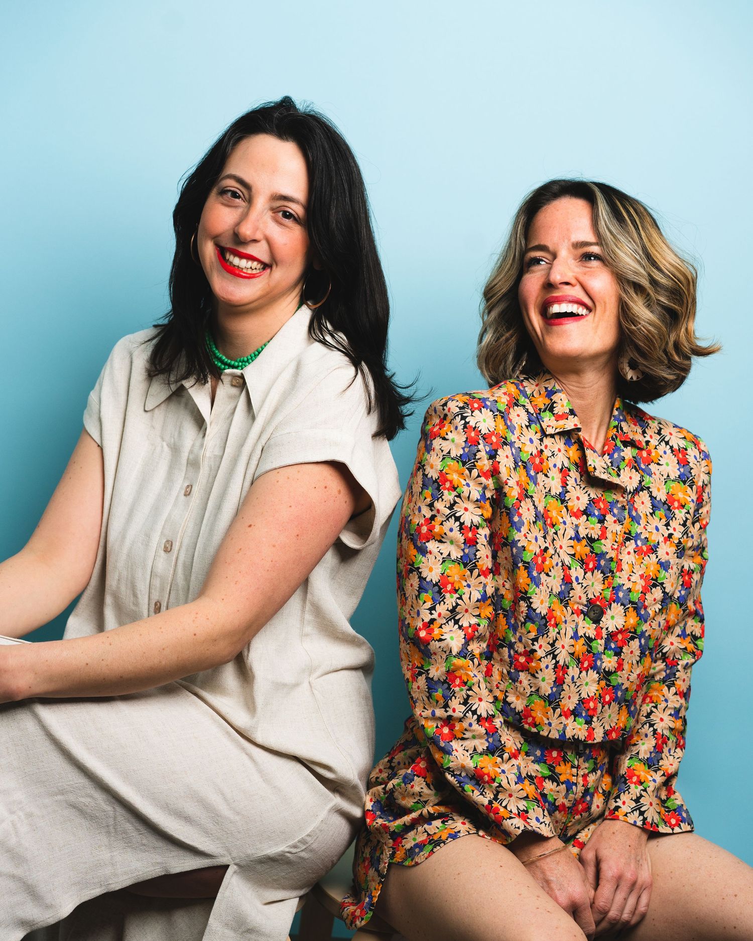 Two people pose together playfully in contrasting outfits of white and floral print against a blue background.