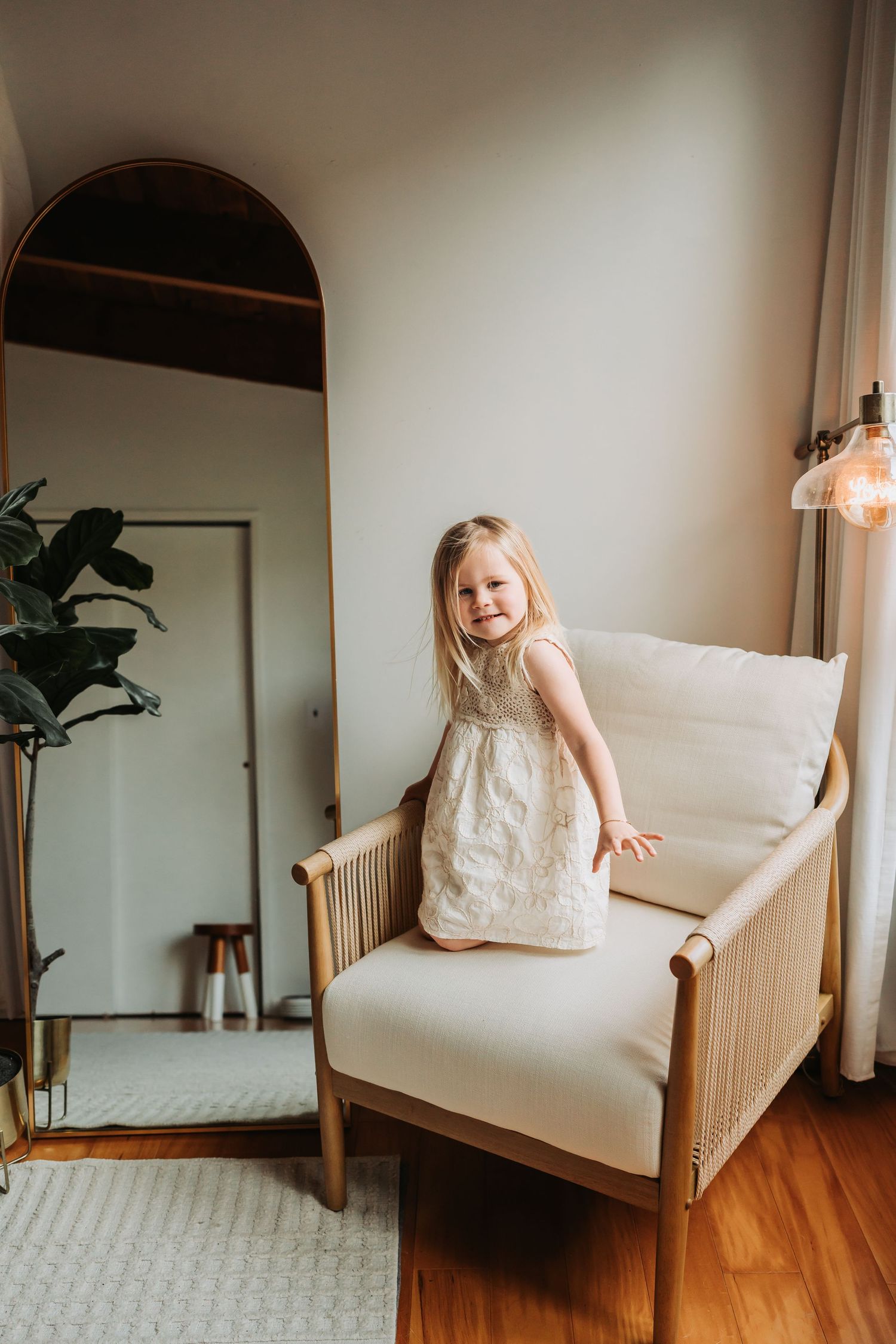Smiling blonde toddler girl in cream dress standing on a white armchair in a bright modern living room.