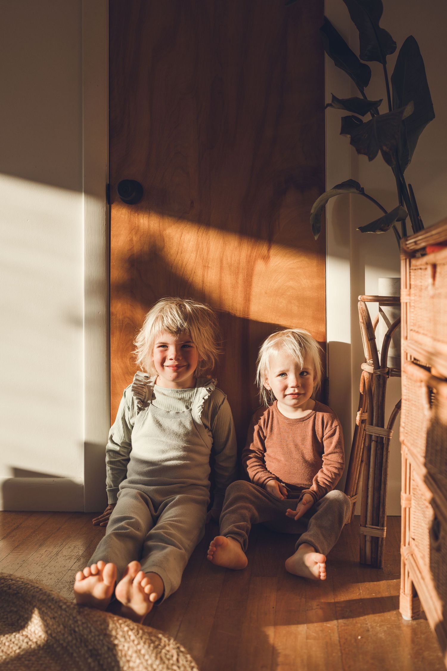 Two young children sit on wooden floor in warm sunlight streaming through window.