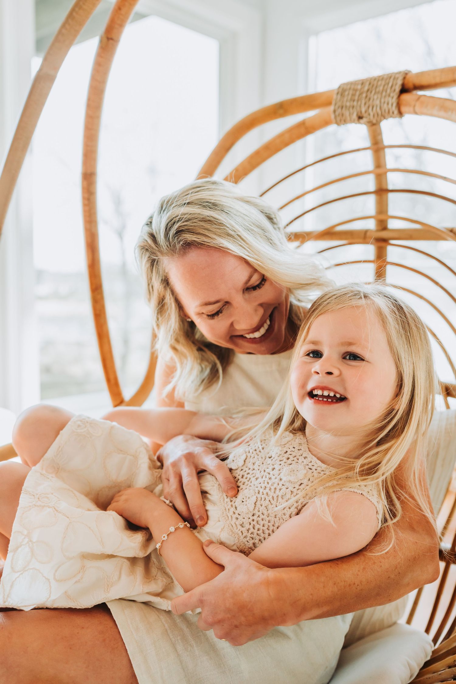 Smiling blonde mother and toddler daughter laughing together in a rattan hanging chair.