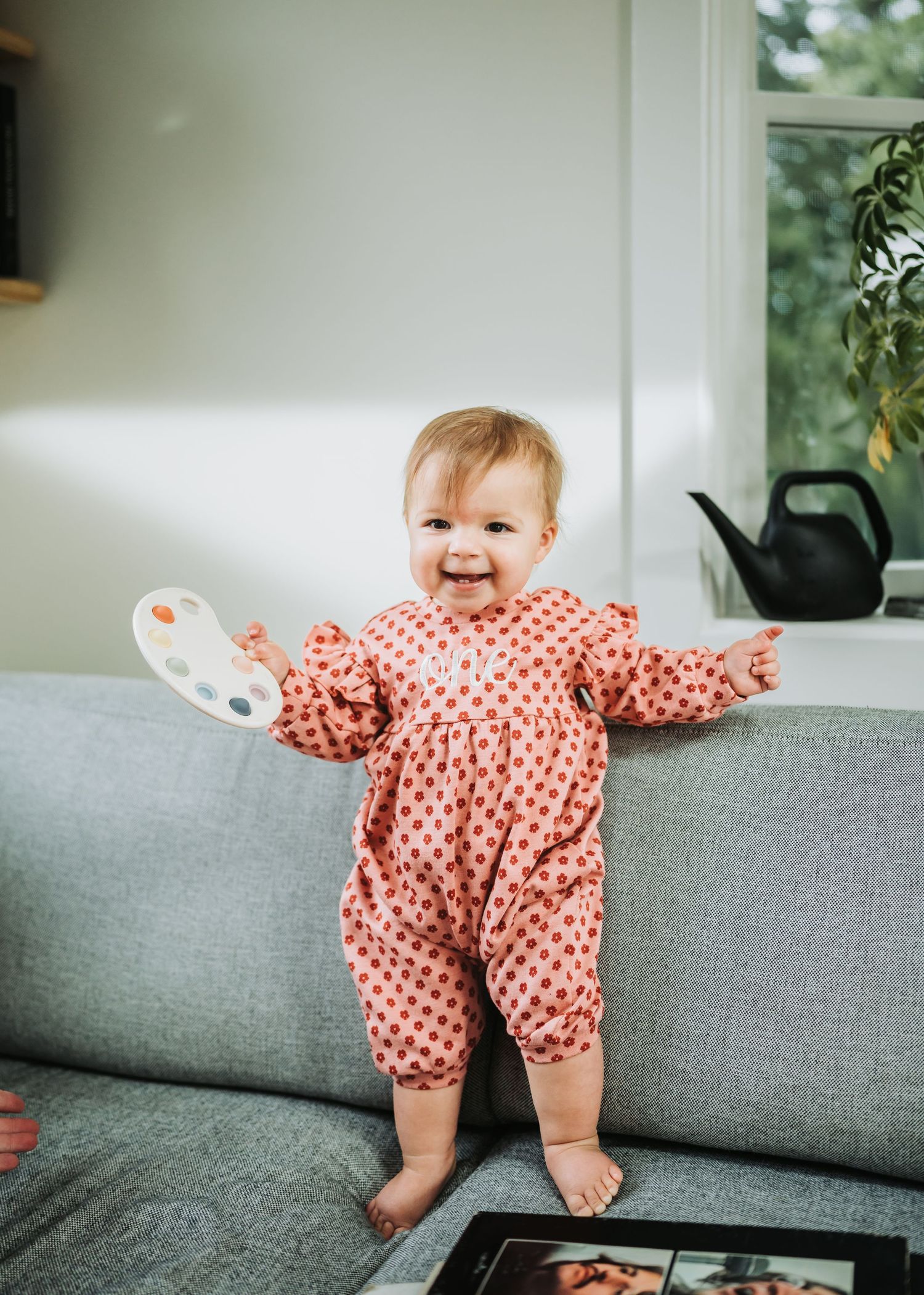Baby in red polka dot romper plays and moves along grey couch in sequential action shots.