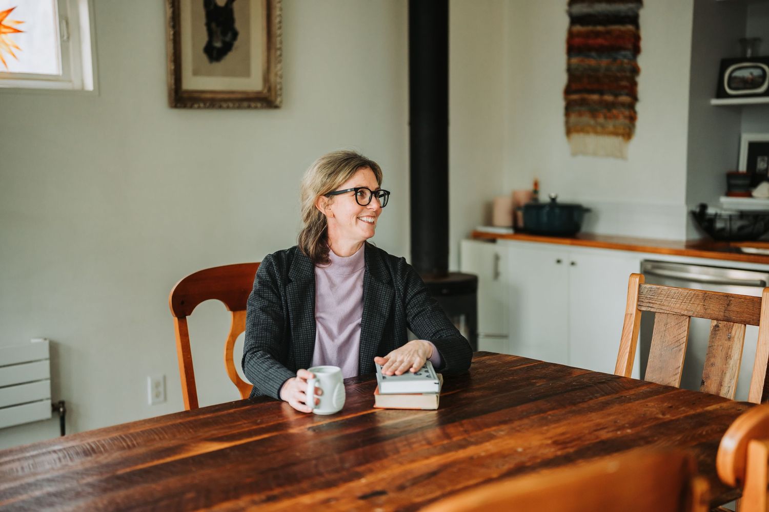 Someone wearing glasses and a dark blazer sits at a wooden dining table with a coffee mug in a bright interior space.