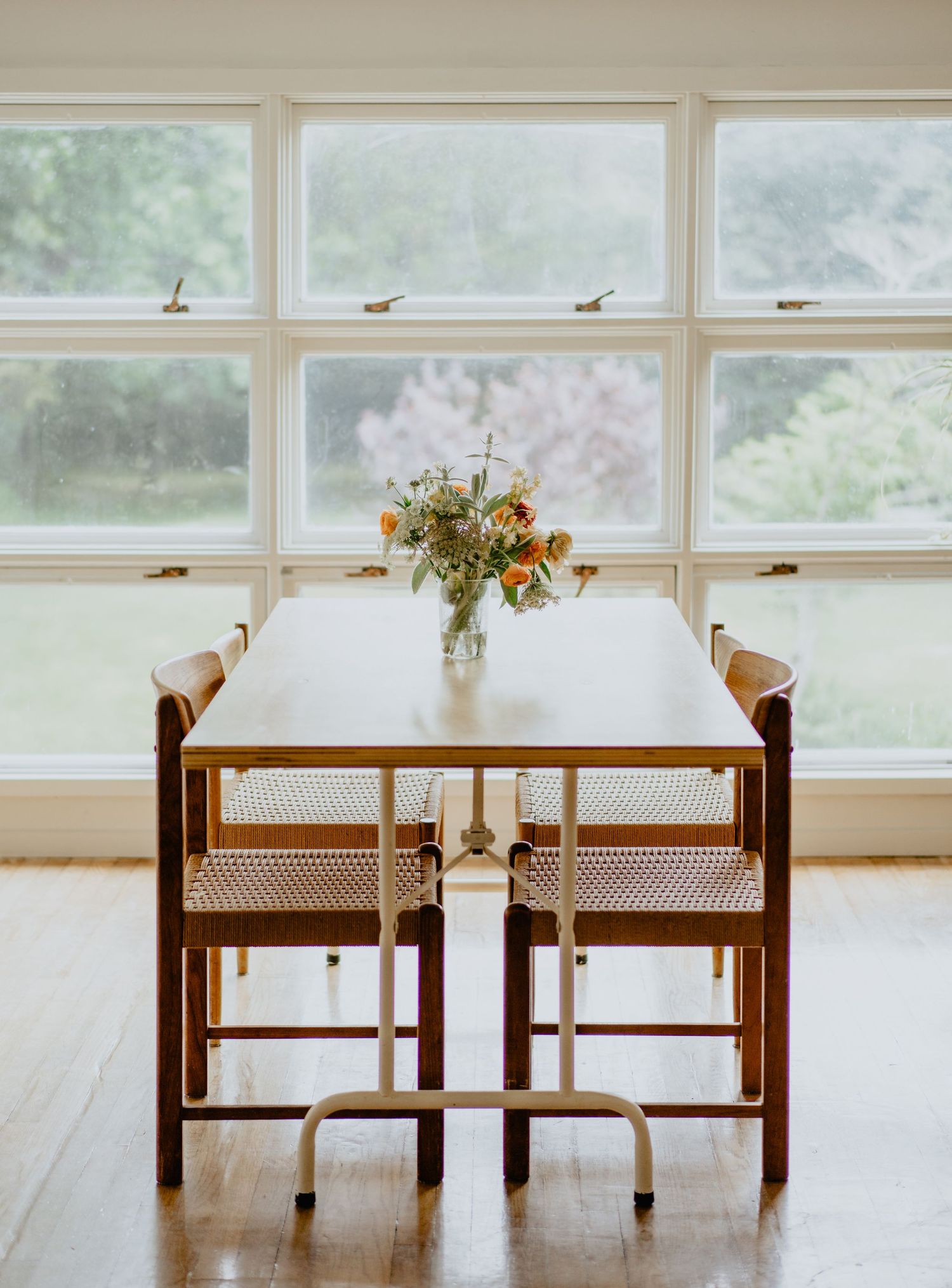 A modern white dining table paired with two wooden chairs featuring woven cane seats next to bright windows.