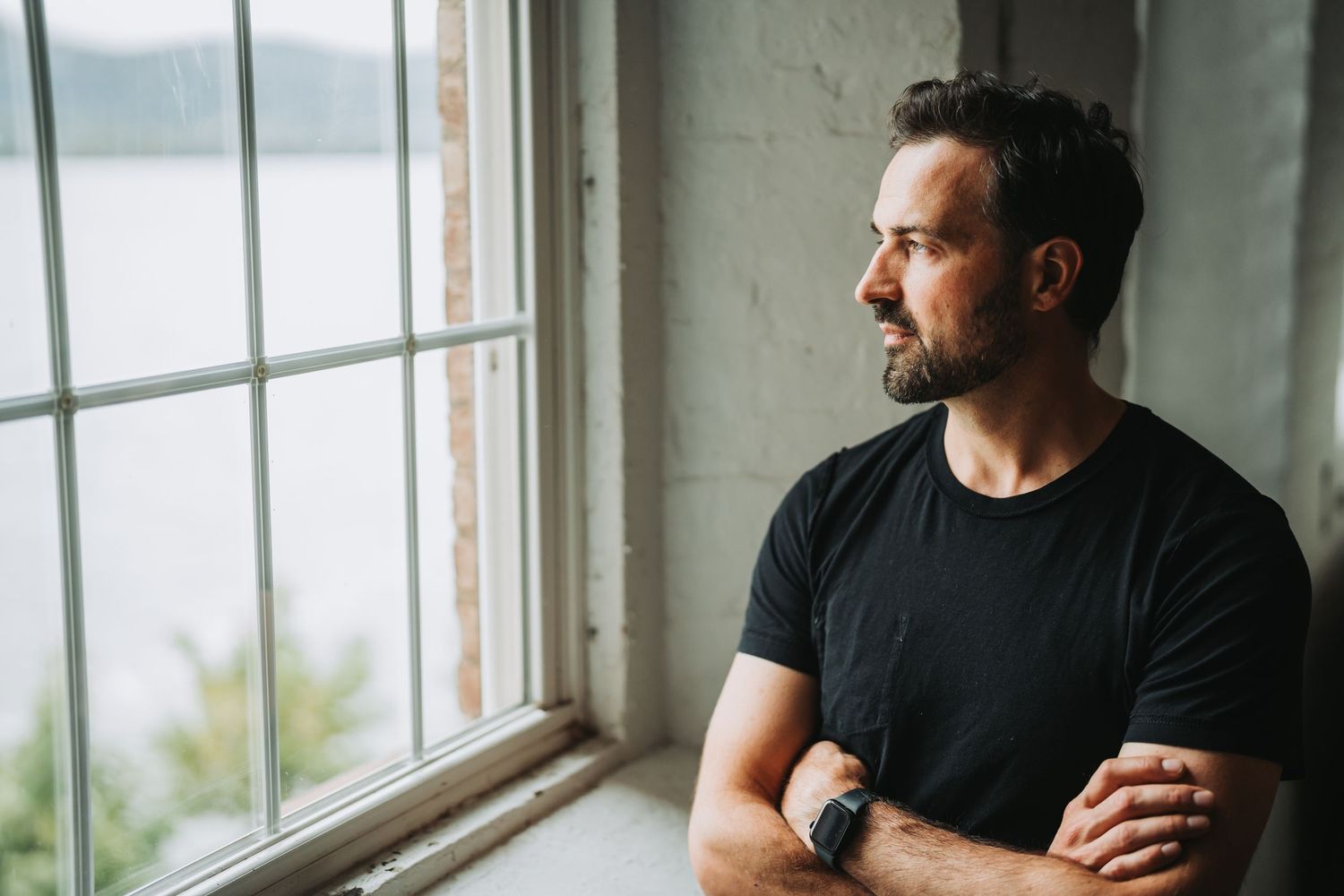 Person in black t-shirt stands contemplatively by window with arms crossed looking at lake view outside.