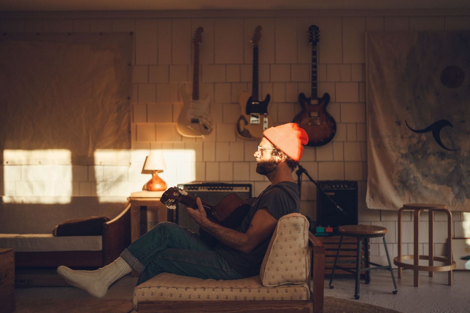Person wearing orange beanie relaxes on couch playing guitar in sunlit room with instruments on wall.