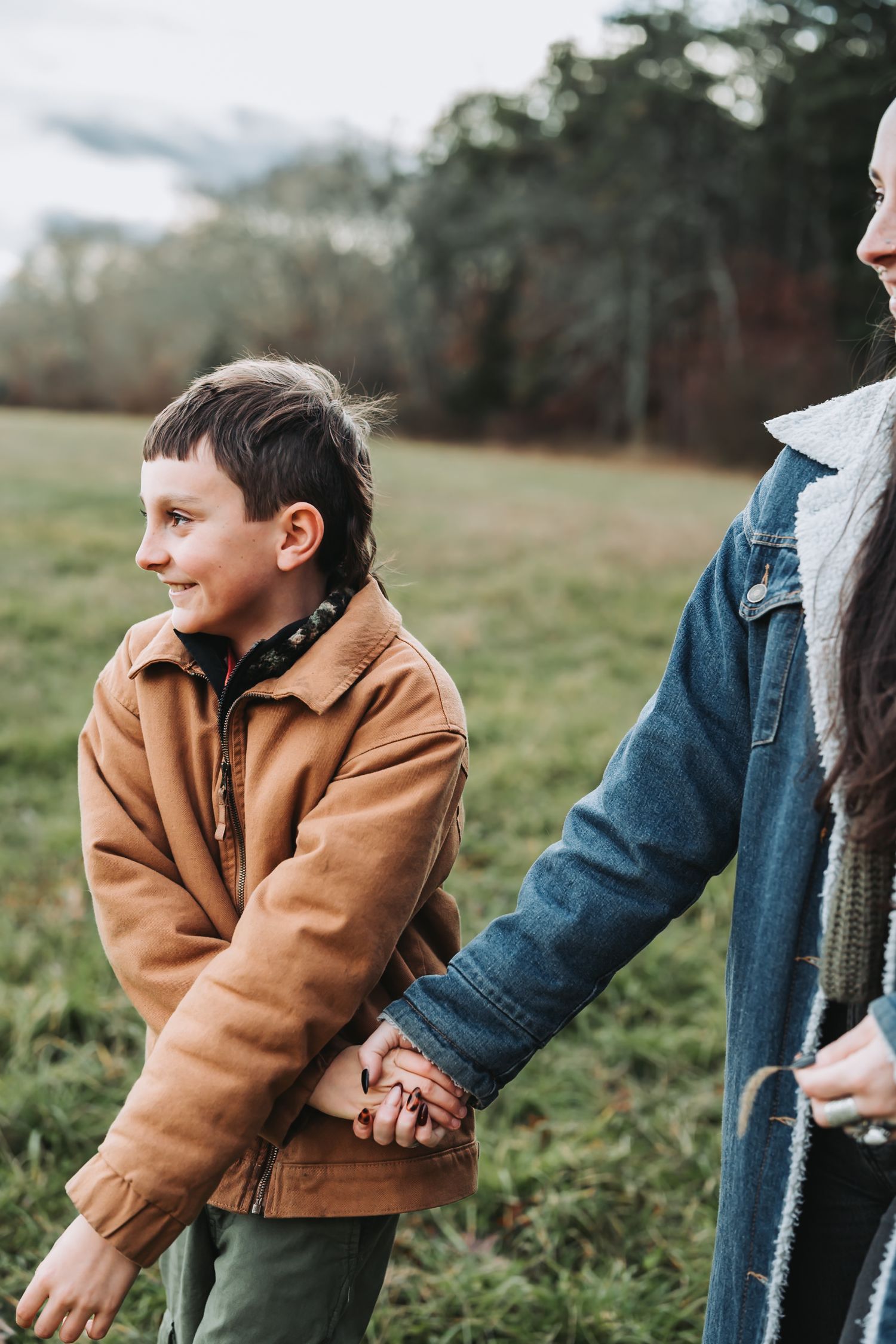 Two people holding hands walk together in an autumn field wearing cozy jackets on a misty afternoon.